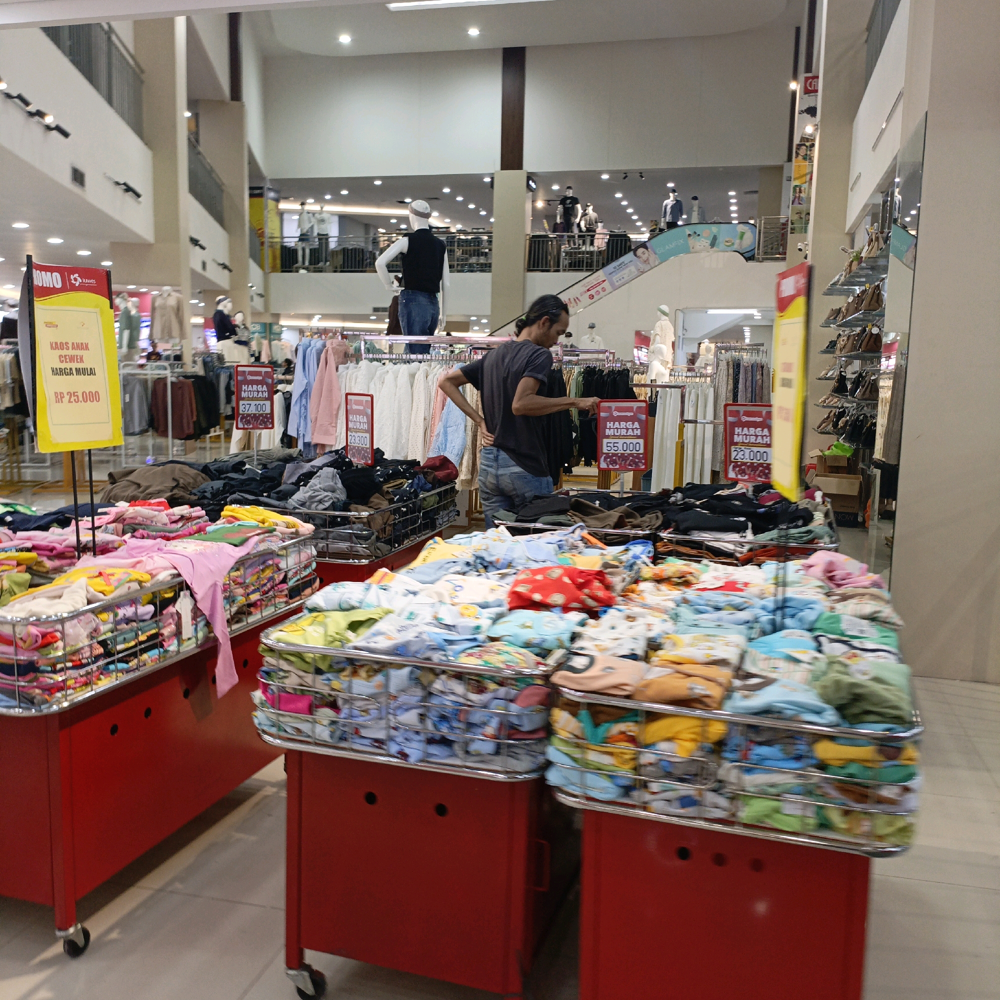 The image shows the interior of a department store in Salatiga, Indonesia. In the foreground, there are several red wheeled display stands filled with folded clothing, primarily children's apparel, some of which is patterned. A person, identified as a male with long dark hair tied back, is visible in the mid-ground wearing a dark t-shirt and blue jeans. He is standing near clothing racks and appears to be looking at items or interacting with a display. In the background, mannequins wearing clothing are displayed on multiple levels of the store, along with rows of hanging garments. Several signs with pricing information are also visible, indicating sales or price points. The overall environment is brightly lit, suggesting a commercial retail space.