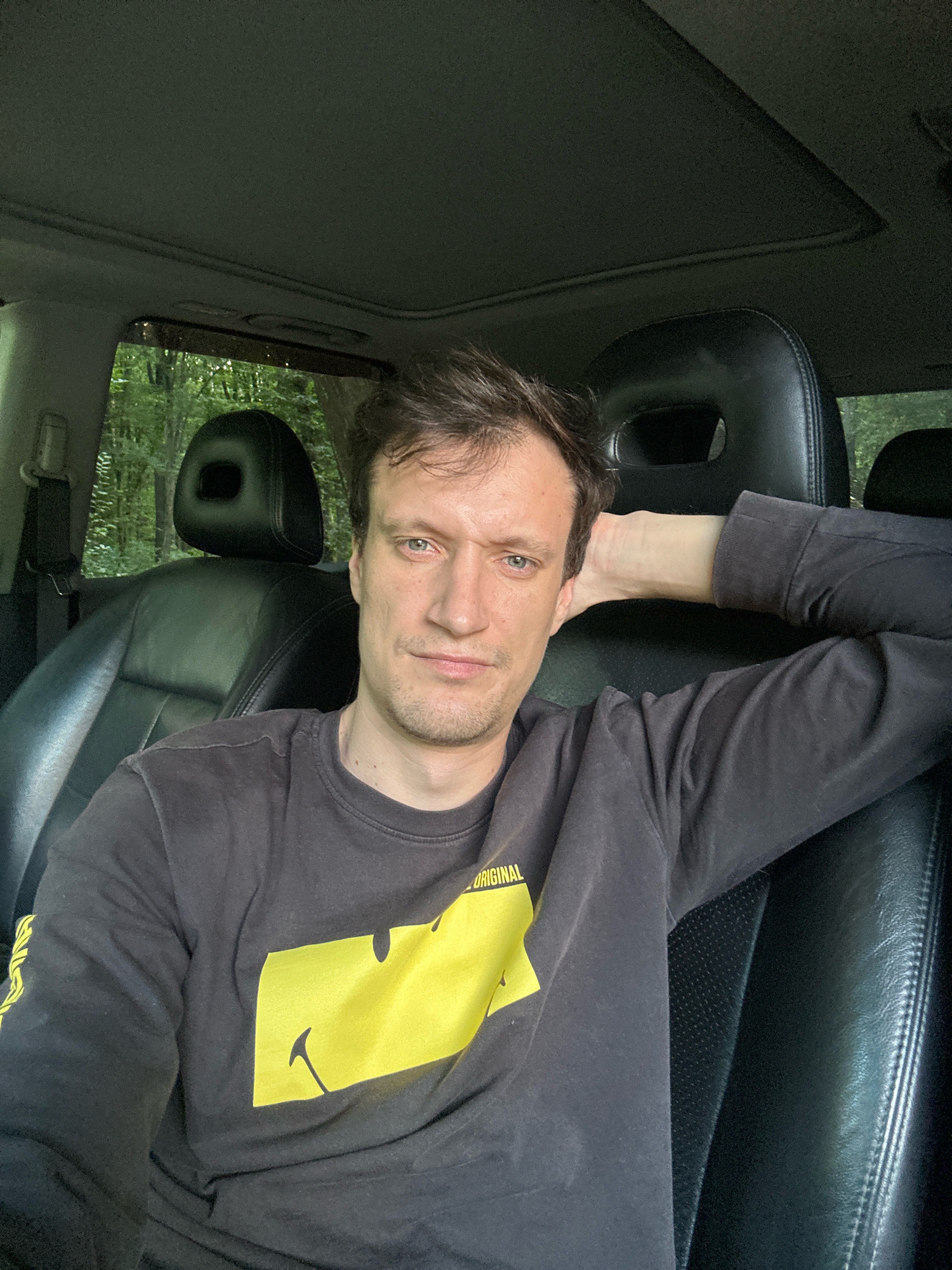 The image is a close-up selfie of a man sitting in the driver's seat of a vehicle. He is wearing a dark long-sleeved shirt with a yellow graphic print. His right arm is resting behind his head, and he is looking directly at the camera with a neutral expression. The interior of the car is visible, including black leather seats and the sunroof. Through the car windows, a lush green forest can be seen. The man's age appears to be between 30 and 40 years old. The location is identified as being in Shatava, Ukraine.