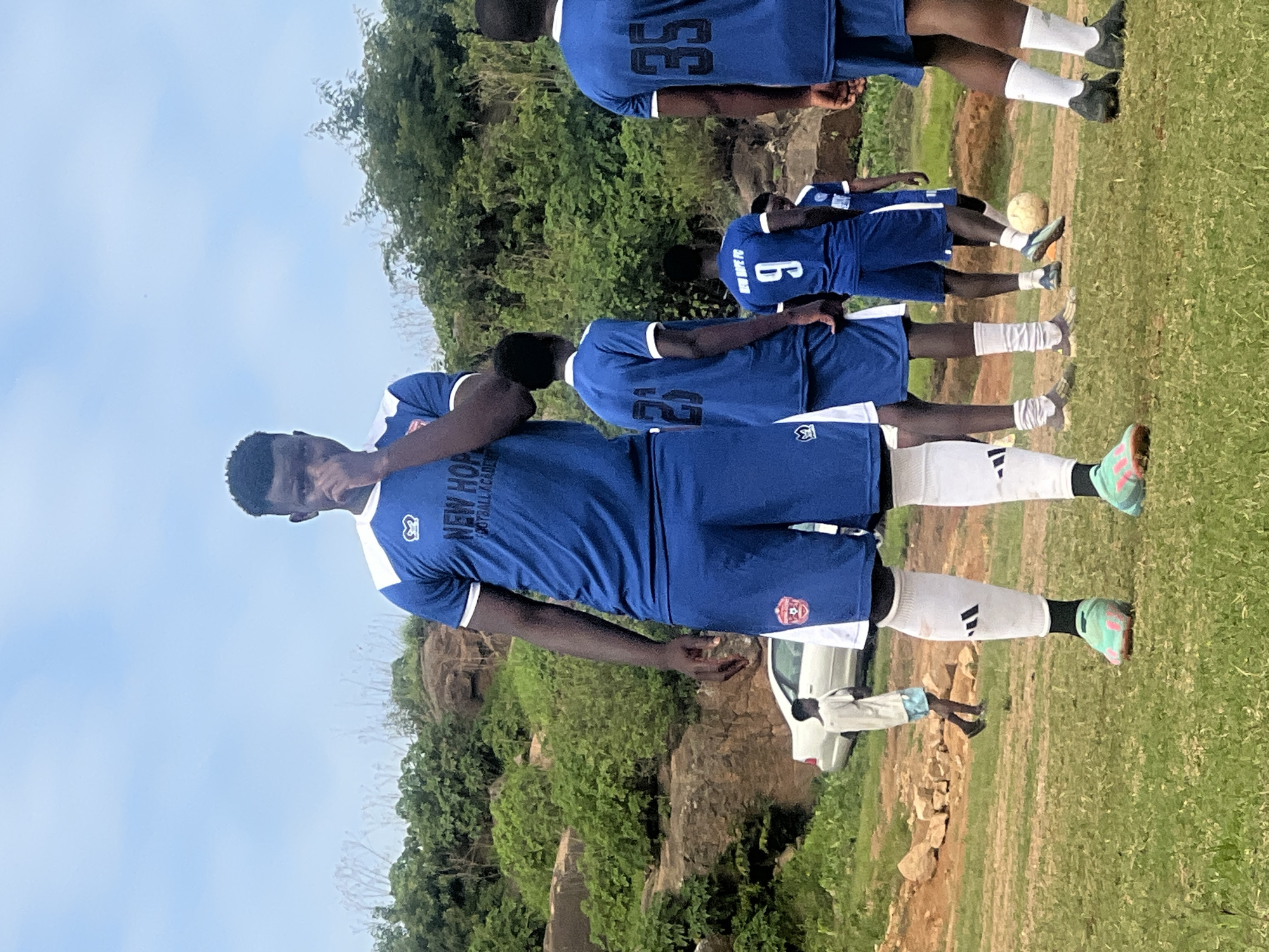 A group of young men, dressed in blue soccer uniforms with white accents, stand on a grassy field. The man in the foreground, closest to the camera, is wearing a blue jersey with "NEW HOPE FOOTBALL ACADEMY" printed on it. He is also wearing blue shorts and white socks. Other players in the background are also wearing similar uniforms, with numbers visible on some of their jerseys, such as "35" and "6". They appear to be a soccer team preparing for or participating in a training session or game.
The scene is outdoors, with a lush green hillside or rocky outcrop in the background, covered in vegetation. The sky is a clear blue. A white car is partially visible on the field behind some of the players. The lighting suggests it is daytime, likely in the morning or afternoon, with no strong shadows indicating harsh sunlight. The overall mood seems to be focused and ready, typical of a sports team. The location is identified as Jos, Nigeria.