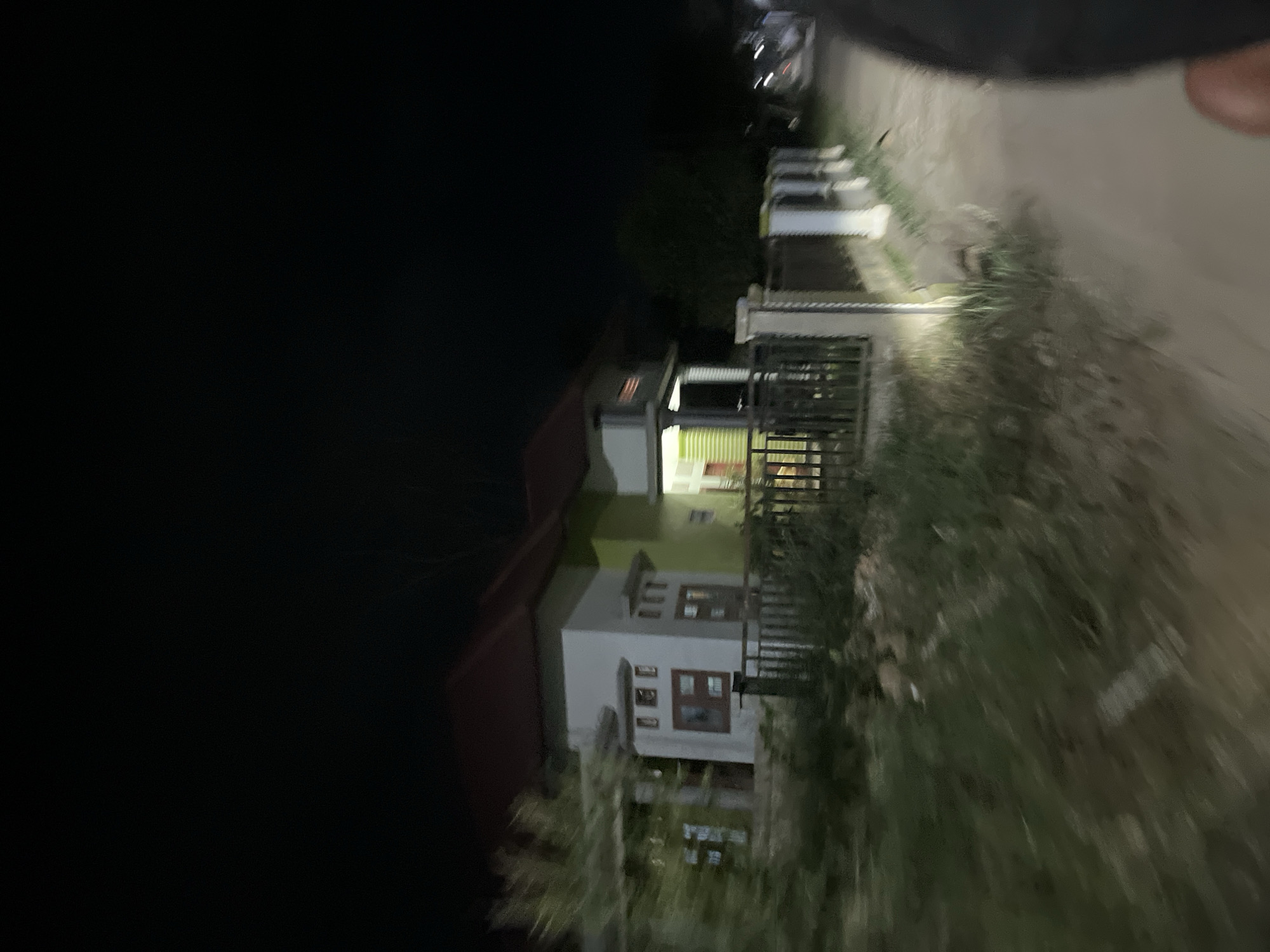 The image shows a residential building at night in Menanggini, Indonesia. The building appears to be two stories tall with a dark-colored roof and white walls. There are multiple windows visible, some with wooden frames. The building is surrounded by vegetation, including trees and bushes. A fence or railing is also visible in front of the building. The scene is dimly lit, suggesting it is nighttime. The image is slightly blurry and appears to be taken from a moving vehicle, indicated by the dark, out-of-focus area in the upper left corner and the tilted perspective. There is no visible text in the image. The overall mood is quiet and residential.