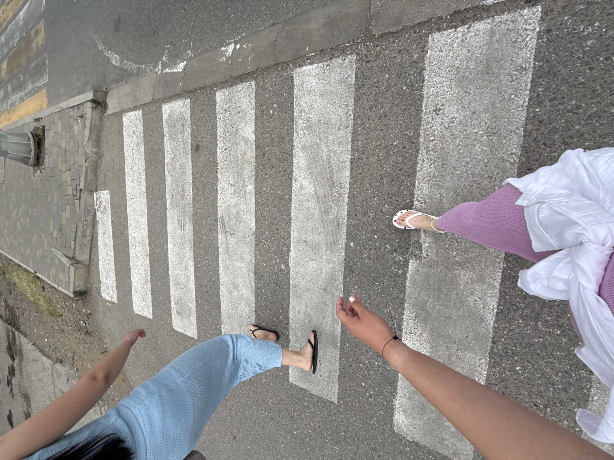 This is a high-angle, first-person perspective shot of two individuals crossing a pedestrian crosswalk. The scene is set on a paved road with clearly marked white stripes for the crosswalk. To the left of the crosswalk, a sidewalk made of interlocking grey pavers is visible, with some sparse green vegetation growing through the cracks. A metal street lamp post is also present on the left side of the frame.
One person, closer to the viewer, is wearing light blue pants and black flip-flops. Their arm is extended to the right, with a black bracelet on their wrist. The other person, further into the crosswalk, is wearing a lilac-colored skirt or pants and white flip-flops with black polka dots. They are also wearing a white garment, possibly a shirt or dress, draped over their shoulder.
The lighting suggests it is daytime, with shadows cast across the crosswalk. The weather appears to be overcast, as there are no harsh shadows. The setting of Derbent, Russia, is provided as context but not visually identifiable from the image itself. No visible text is present in the media file. The activity is simply crossing the street, and the overall mood is casual.