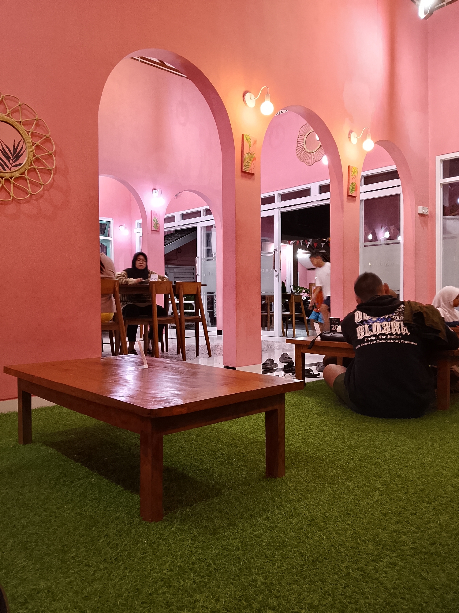 The image depicts the interior of a cafe in Mernung, Indonesia. The dominant color scheme is pink, with pink walls and arched doorways creating a warm and inviting atmosphere. The floor is covered in artificial green grass, adding a unique touch to the decor.

In the foreground, a wooden coffee table sits on the grass. In the background, several people are visible. One woman is seated at a table with chairs, wearing glasses and a patterned top. Another person, a man wearing a black hoodie, is seated on the floor facing away from the camera. Further in the background, a person in a white shirt and blue shorts is walking. Several other individuals are partially visible in the distance.

Decorative elements include a woven mirror with a plant design on the left wall and two framed pictures with botanical motifs hanging on the walls near the arches. Lights are mounted on the walls above the arches, providing illumination. The cafe appears to be open with large windows and doors leading outside, suggesting an indoor-outdoor seating arrangement. The overall impression is a casual and trendy cafe environment.