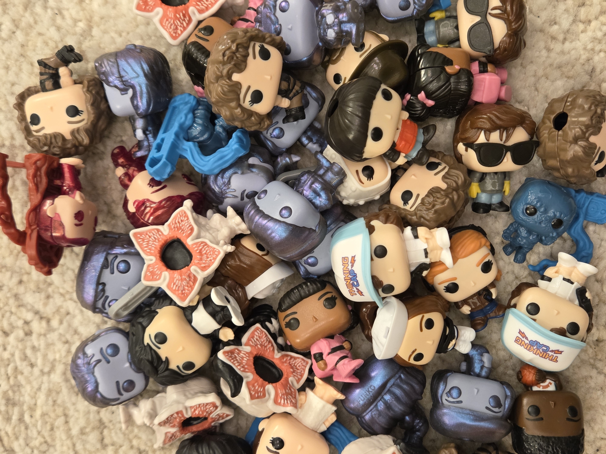 A close-up, overhead shot depicts a disorganized pile of miniature Funko Pop figurines. The figures are mostly head and torso shots, with no clear arrangement. The subjects of the figures are varied, including humanoids, aliens, and creatures. The figures are made of plastic, and most of them are in the color scheme of brown, blue, or purple. The background appears to be a cream-colored carpet or rug. The location is not depicted in the image.