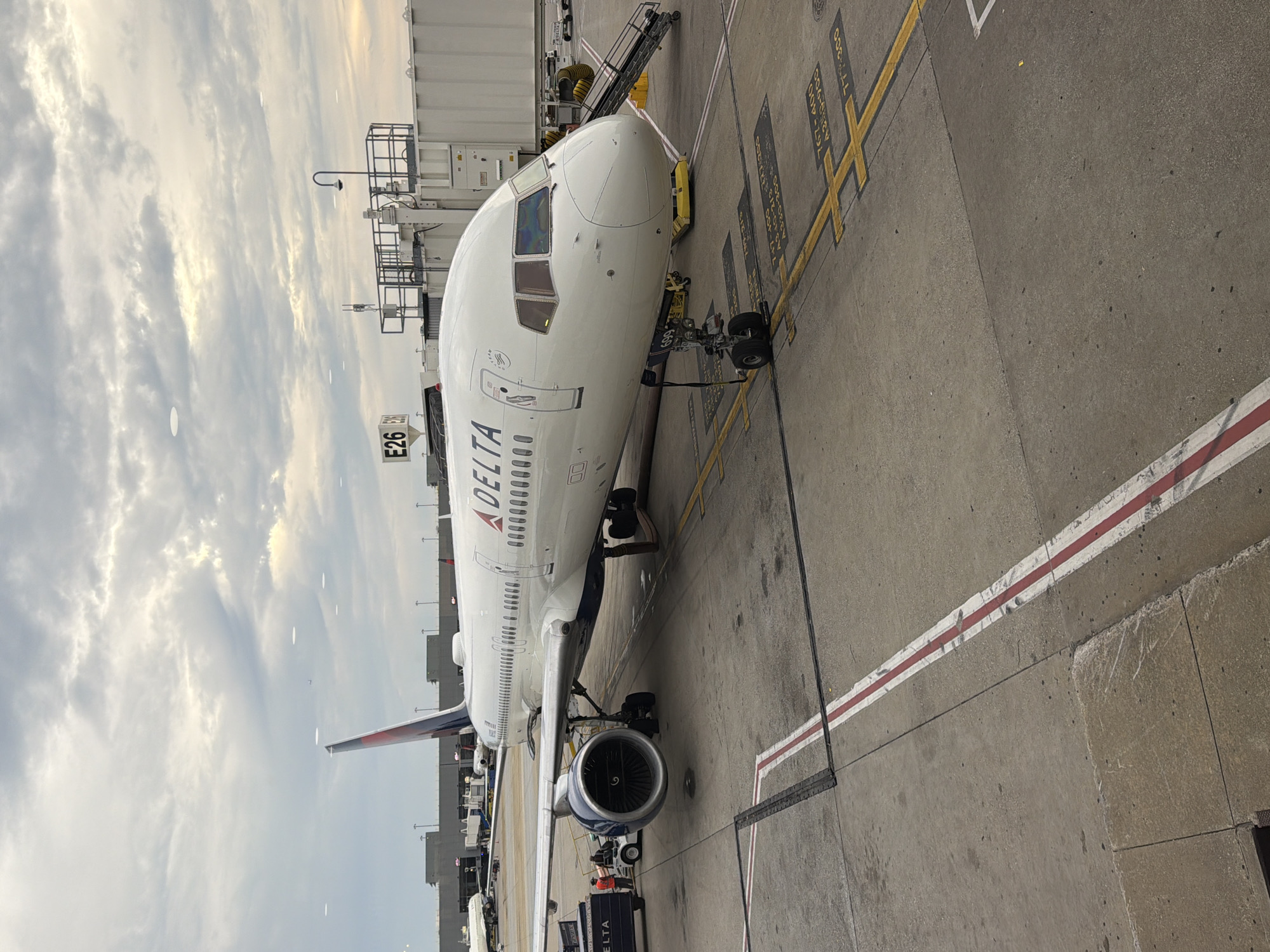 A Delta Airlines airplane is parked at Gate E26 at Hartsfield-Jackson Atlanta International Airport. The aircraft, a Boeing 757-200, is white with red and blue Delta branding. It is connected to a jet bridge, and ground crew are visible near the front landing gear and to the left of the plane. The tarmac is marked with yellow lines and painted instructions for aircraft parking. The sky above is overcast with scattered clouds. The image captures a typical scene at a major airport.