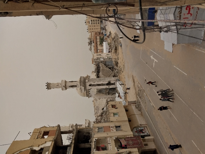 The image captures an elevated view of a heavily damaged urban street scene, likely after conflict or a natural disaster. The perspective is from an upper floor of a building, looking down onto the street and surrounding structures.
In the immediate foreground, what appears to be the edge of a building and some overhead utility wires (electrical or communication) are visible, partially framing the top of the scene.
The mid-ground is dominated by widespread destruction. On the left, several buildings are severely damaged, with walls collapsed, interiors exposed, and rubble scattered around. A prominent feature in this area is a mosque, identifiable by its white minaret and dome, which also shows signs of damage but remains largely intact. More destroyed buildings surround the mosque.
A wide, paved road runs through the center and right side of the frame. White markings are visible on the asphalt, including a dashed center line and a large white arrow pointing away from the viewer, indicating traffic flow. Scattered debris and dirt are visible along the edges of the road.
Several people are present on the street. Towards the foreground of the road, a person dressed in dark clothing (possibly with a red element) is interacting with a soccer ball, suggesting play or casual activity amidst the ruins. Further up the street, a small group of about five individuals, also in dark clothing, are gathered together. In the distance, near the top right of the frame, two more figures are walking along the street.
In the background, beyond the immediate destruction, some buildings appear relatively less damaged, suggesting parts of the city that may have been less affected. The sky is a uniform, light, hazy grey-brown, indicating either an overcast day or the presence of dust or smoke in the air.
Visible text includes Arabic script on a large white banner or sign located on the right side of the street, near the middle-ground. A prominent red logo, possibly a stylized human figure or symbol, is also on this banner. Parts of the Arabic text are legible, including "مؤسسة" (mu'assasah - institution/foundation) and "مطعم" (mat'am - restaurant), followed by other words like "على" (ala - on/upon). A smaller red sign with some text is also visible on a building to the left of the road, but it is too small to decipher clearly.
The scene conveys a sense of desolation and the impact of widespread damage, yet also hints at a continuation of daily life with people, including children, on the street.