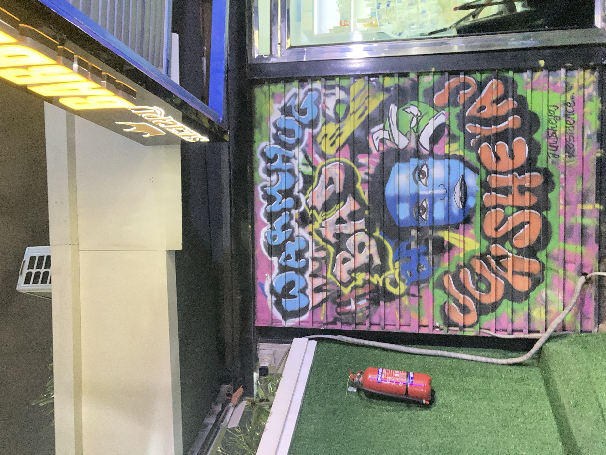 The image shows a close-up of a graffiti-covered rolling shutter door on what appears to be a shopfront. The graffiti features a vibrant blue face with dark eyes and lips, layered over colorful abstract shapes and text. The text appears to be in English, with some parts legible like "WARMTHOGS," "BAD," and "SHEILA." Below the graffiti, a small signature or tag reads "juusheyej residence...". To the left of the shutter, a red fire extinguisher is mounted on a green textured wall. Above the shutter, a white awning is visible, and a sign with yellow illuminated lettering, partially obscured, reads "BARB..." suggesting it might be a barbershop. The scene is likely set in Abuja, Nigeria, given the provided context. The lighting suggests it is evening or night.