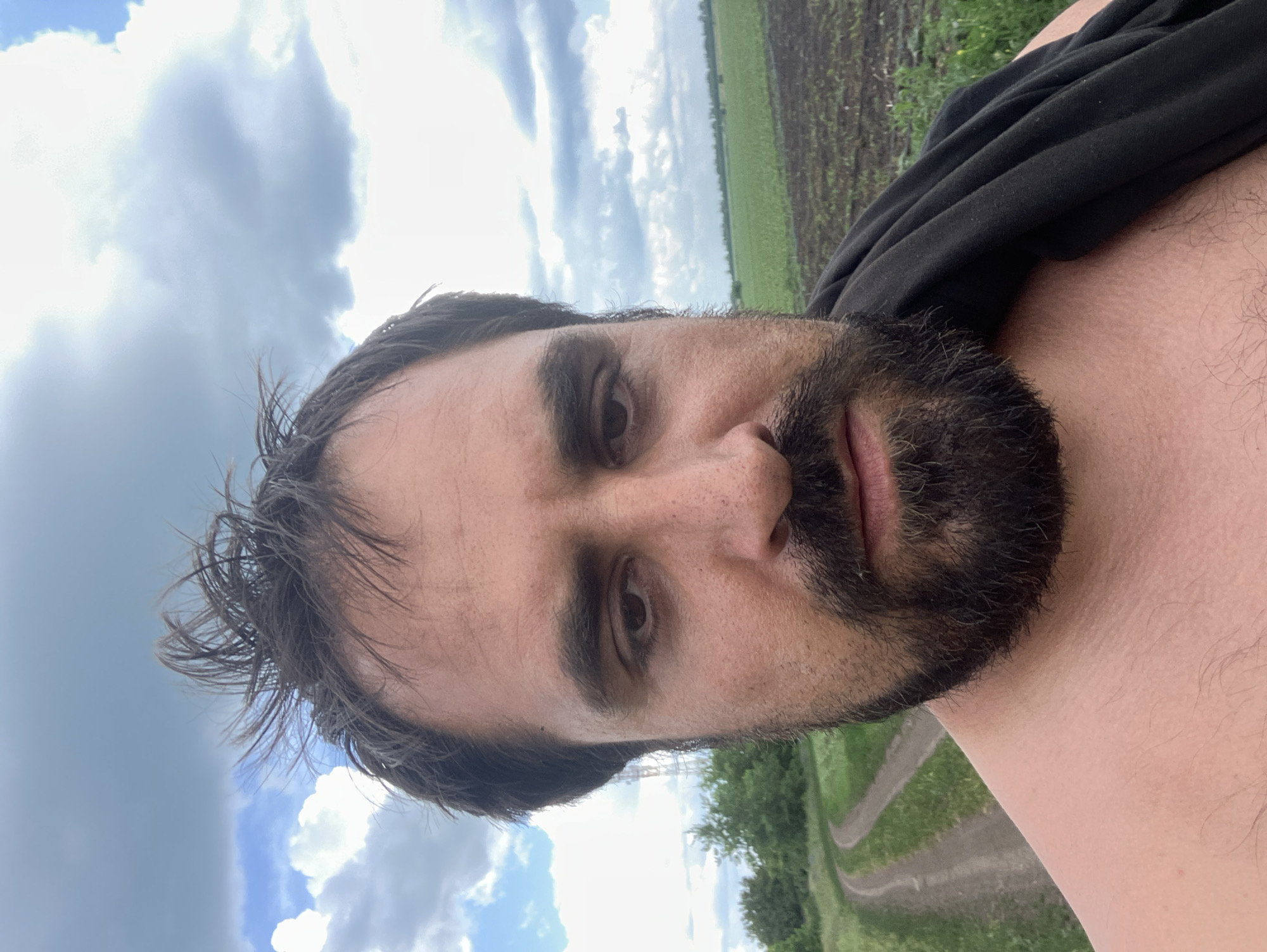 The image is a close-up selfie taken outdoors in Berdiansk, Ukraine. The subject is a man with dark hair, a beard, and fair skin, looking directly at the camera. He is shirtless, with a black garment draped over his right shoulder. The background features a cloudy sky, a dirt road curving through a grassy field, and a cultivated field in the distance. The overall scene suggests a rural or semi-rural environment.