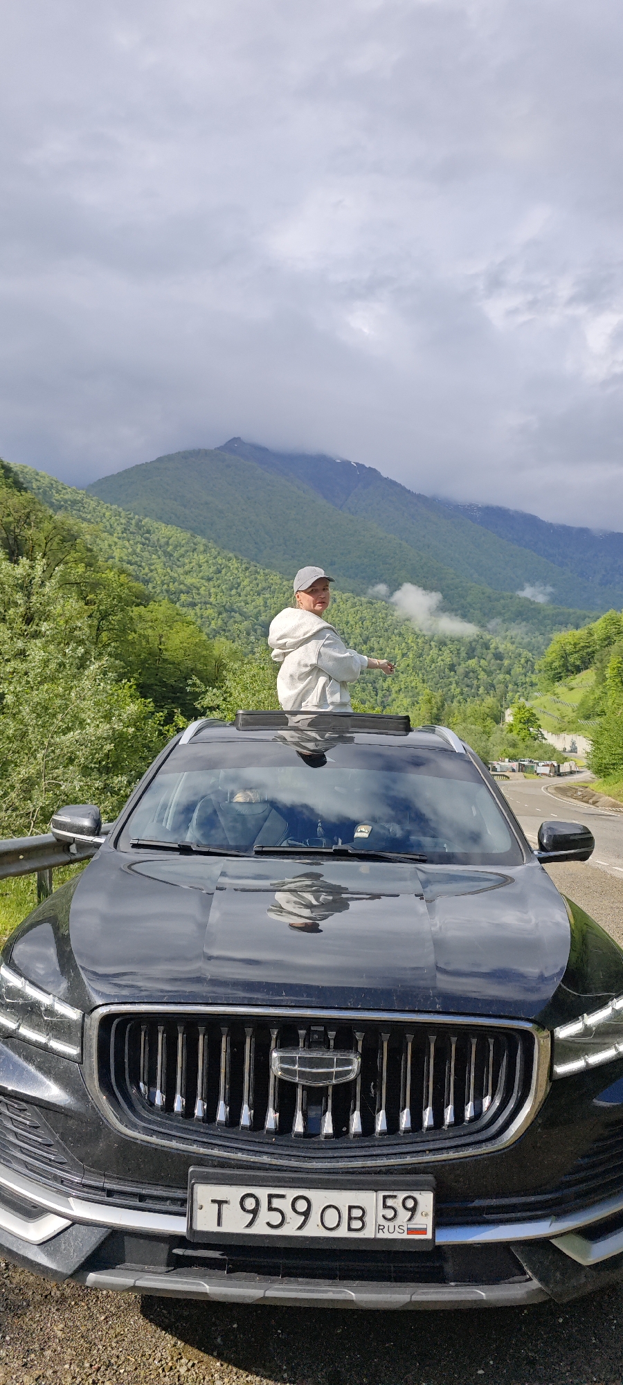 A medium shot presents a black car parked on a roadside with a person standing through the open sunroof. The person, wearing a light-colored hooded sweatshirt and a hat, stands upright, their body facing away from the camera and their right arm outstretched, as if pointing at the scenery.
The car faces the camera directly, and its front grill and license plate reading "T 959 OB 59 RUS" are clearly visible. The car's hood reflects the surrounding sky and the person standing above.
The background consists of a mountainous landscape covered with green trees. The mountain peaks in the distance are partially obscured by clouds. The road curves to the right, and a line of parked vehicles can be seen in the distance along the roadside.
The sky is overcast with gray clouds. The environment suggests a rural or mountainous area.