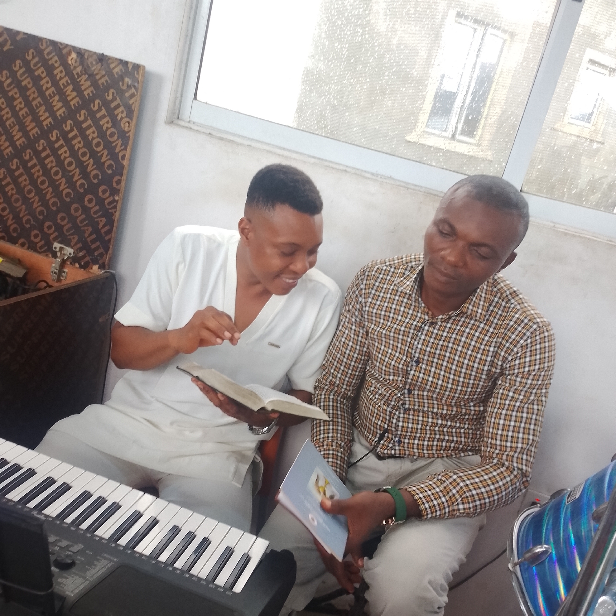 In Iranla, Nigeria, two men are seated indoors, possibly in a classroom or community center. One man, wearing a white tunic, holds an open book and smiles as he looks at the other man. The second man, dressed in a plaid shirt and white pants, looks down at a book in his hands. To the left of the frame, a black keyboard and a wooden board with "SUPREME STRONG QUALITY" printed repeatedly are visible. On the right, a blue drum set with a chrome finish is partially visible. The environment appears to be brightly lit by natural light filtering through a large window with a view of buildings.