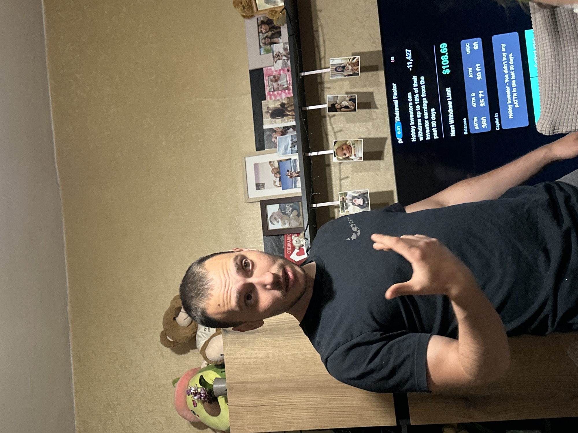 A medium shot captures a man in a dark t-shirt standing in front of a television screen displaying financial information. He has short, dark hair and is gesturing with his right hand. Behind him, on a shelf, are several framed photographs and plush toys, including a teddy bear and an avocado-shaped cushion. The television screen shows text related to "Withdrawal Factor," "Hobby Investors," "Balances," and "Capital In." The room appears to be a living space, with a textured wall and a wooden cabinet. The scene is indoors and appears to be in a residential setting. No specific location in Sofia, Bulgaria, can be determined from the image.