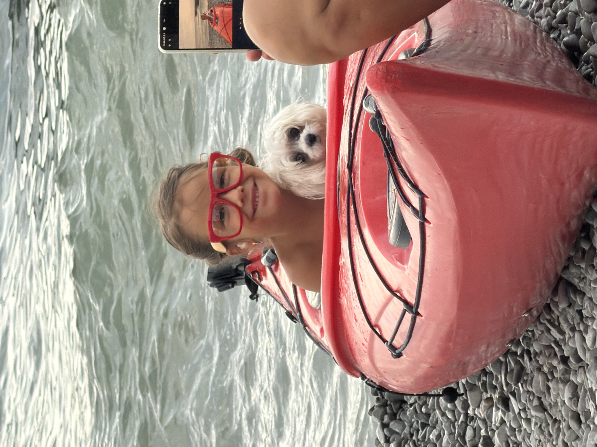 A young girl wearing red glasses and a small white dog are in a red kayak near a pebble beach and a body of water. The girl is smiling and looking at the camera, while the dog is peeking over the edge of the kayak. A person's arm and a smartphone are visible in the foreground on the right, with the phone displaying an image of the girl and dog in the kayak. The scene appears to be at the coast of Partenit, Ukraine.