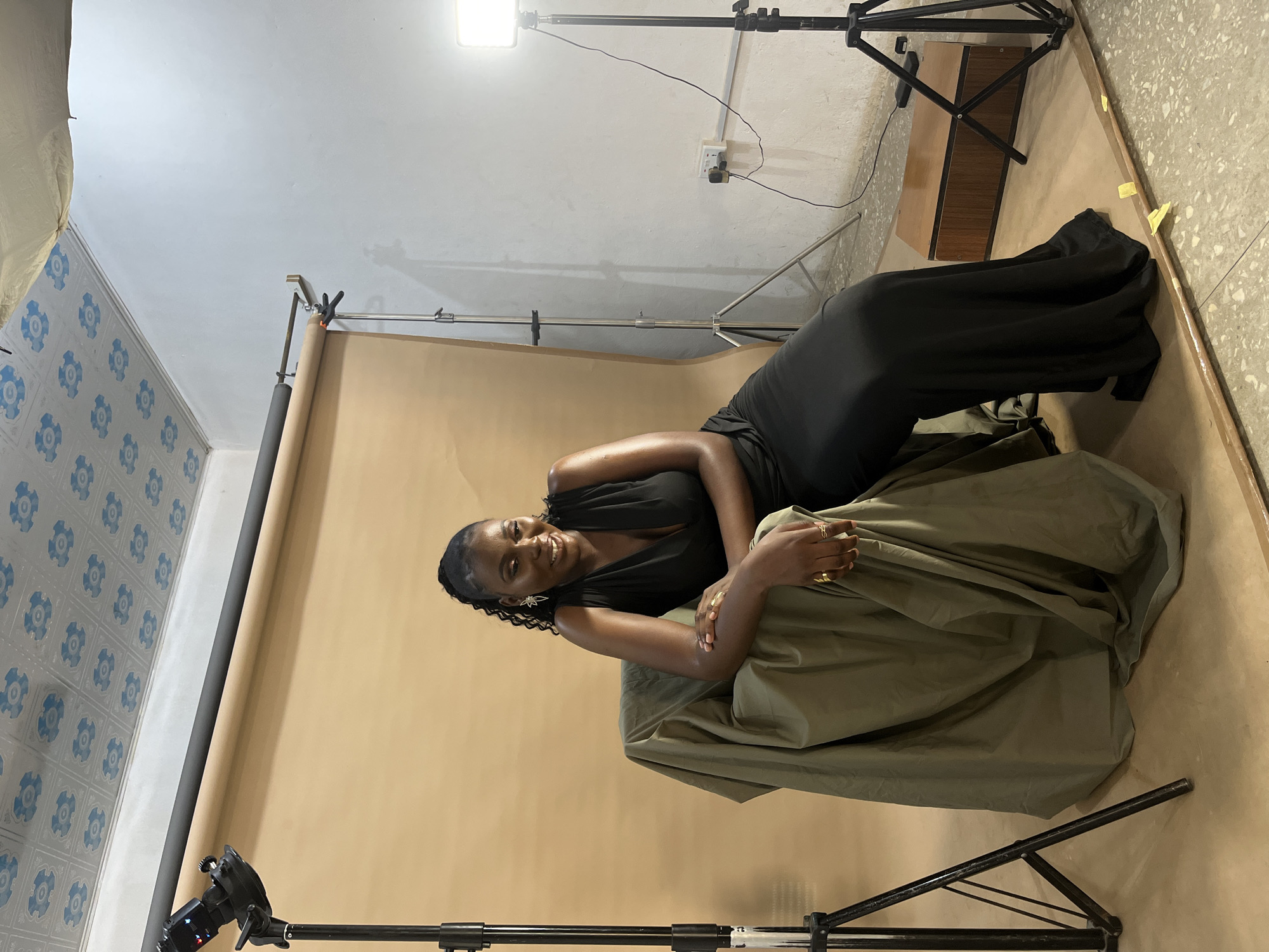 A woman in a black dress is posing for a photoshoot in a studio setting. She is lying down on a draped olive-green fabric, with a beige backdrop behind her. The woman has dark skin and braided hair, and she is smiling at the camera. She is wearing rings on her fingers and star-shaped earrings.
The studio is equipped with professional photography gear, including lighting equipment and camera stands. A large light source is visible in the upper left corner of the image, casting a bright glow. Another light stand is positioned to the right. The walls of the studio are painted white, and one wall is decorated with a blue and white patterned wallpaper. In the background, a wooden cabinet is visible. The floor is concrete.
The overall mood of the image is relaxed and professional, suggesting a creative session taking place. The lighting is bright and even, characteristic of a studio environment. No specific time of day or weather conditions are discernible. The location context provided is Gura Top, Nigeria. There is no visible text in the image.