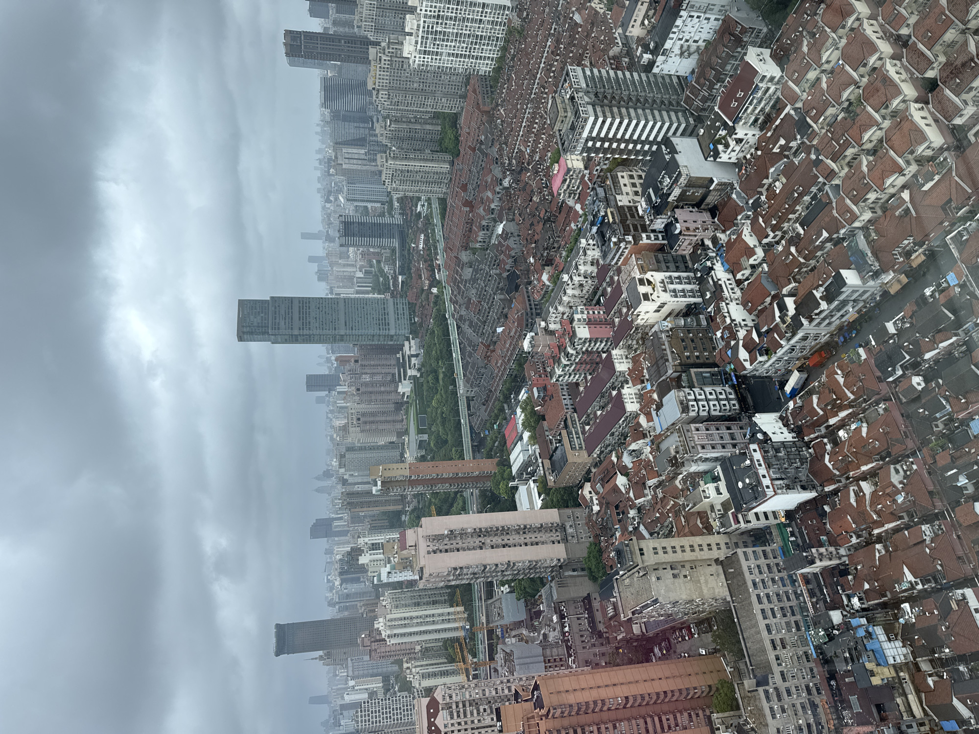 This is an aerial view of a dense urban environment in Shanghai, China, under a cloudy sky. The cityscape is characterized by a mix of architectural styles and heights, dominated by numerous buildings. In the foreground, a large cluster of low-rise buildings with red-tiled roofs creates a distinct pattern, suggesting an older residential or commercial district. These buildings are tightly packed, with narrow streets or alleys running between them.
Moving further into the mid-ground and background, the architecture transitions to taller buildings, including apartment blocks and modern high-rise skyscrapers. Some of these taller structures are visible in the distance, interspersed with the denser, lower-rise areas. The overall impression is one of significant urban development and density. The weather appears overcast, which affects the lighting and the overall mood of the scene, lending it a muted, greyish tone. No people or specific actions are discernible from this aerial perspective. The image captures a vast expanse of the city, showcasing its layered development from older structures to contemporary high-rises.