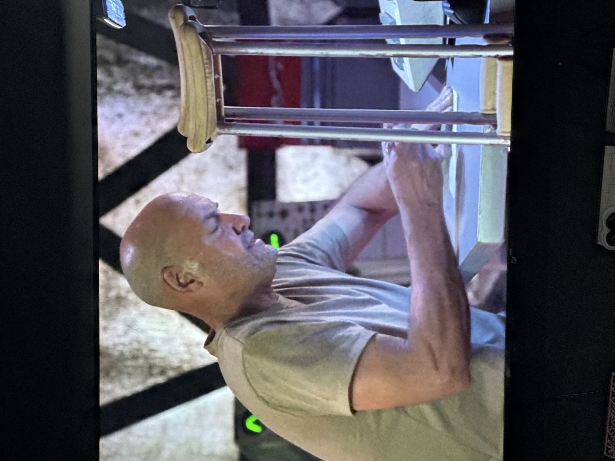 A bald, middle-aged man with a light beard is working at a table. He is wearing a light-colored t-shirt. In front of him is a medical crutch with a yellow padded rest. The background appears to be a room with some industrial elements and what looks like a red metal object. The scene is captured from a medium distance, suggesting it might be a still from a video or television program. The image quality is not high, with some pixelation and a screen-like reflection. There is no indication of the city of Gölbaşı, Turkey, in this image.