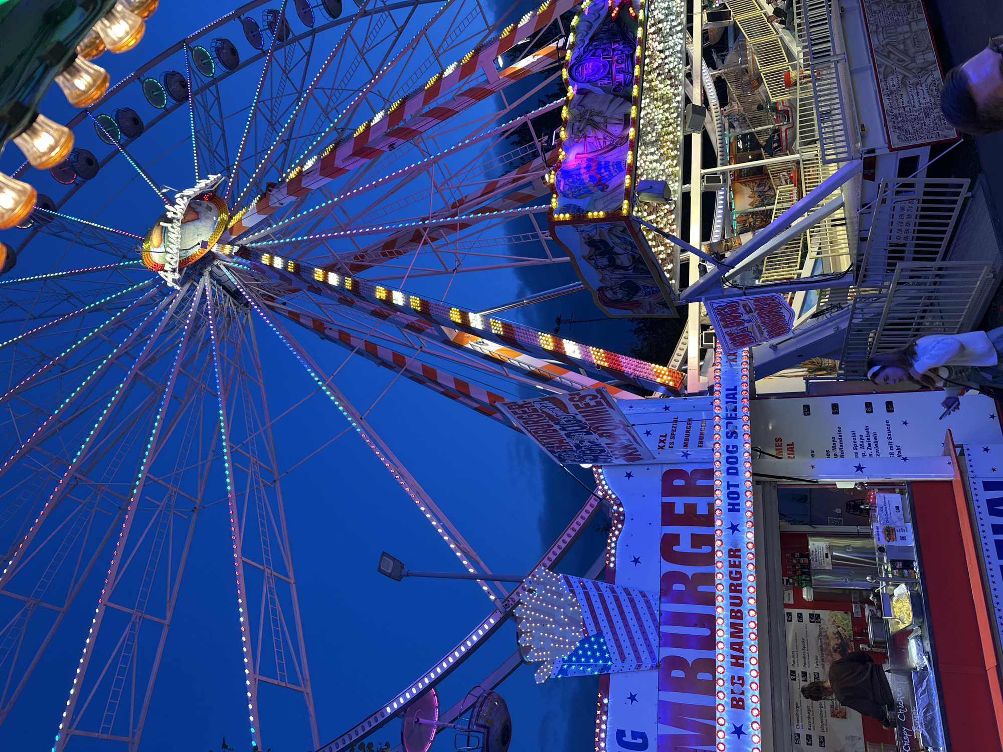 This is a low-angle, eye-level, full shot of a Ferris wheel and food stands at a fair at dusk in Bruchhausen-Vilsen, Germany. The Ferris wheel is the largest object in the frame, with white spokes and a colorful array of lights running along its structure. The sky behind it is a deep blue, transitioning to darker hues with some clouds visible. In the foreground, several food stands are illuminated by strings of lights. One stand prominently displays "BIG HAMBURGER" and "HOT DOG SPECIAL" on a banner. Another stand has a graphic of a box of french fries decorated with the American flag pattern. People are visible at the food stands, with one person in the foreground appearing to order food and another woman standing to the right, looking at her phone. The overall atmosphere suggests a lively evening at a fair.