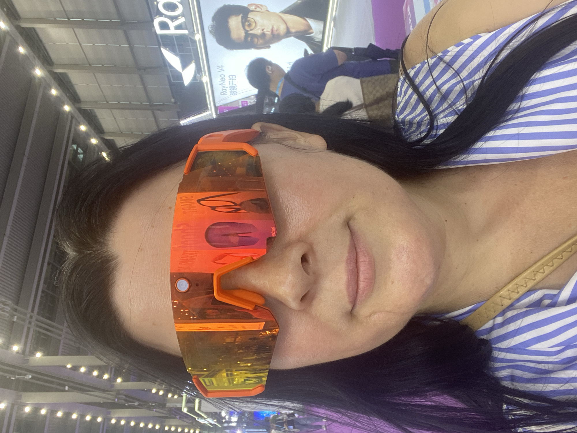 A woman with long dark hair, wearing large orange-tinted wraparound sunglasses and a blue and white striped shirt, is pictured at what appears to be an indoor exhibition or trade show. The background shows an event space with booths, lighting, and a large display screen showing a man wearing glasses and the text "RayNeo V4 眼镜开启". The woman is looking directly at the camera. The scene is brightly lit.