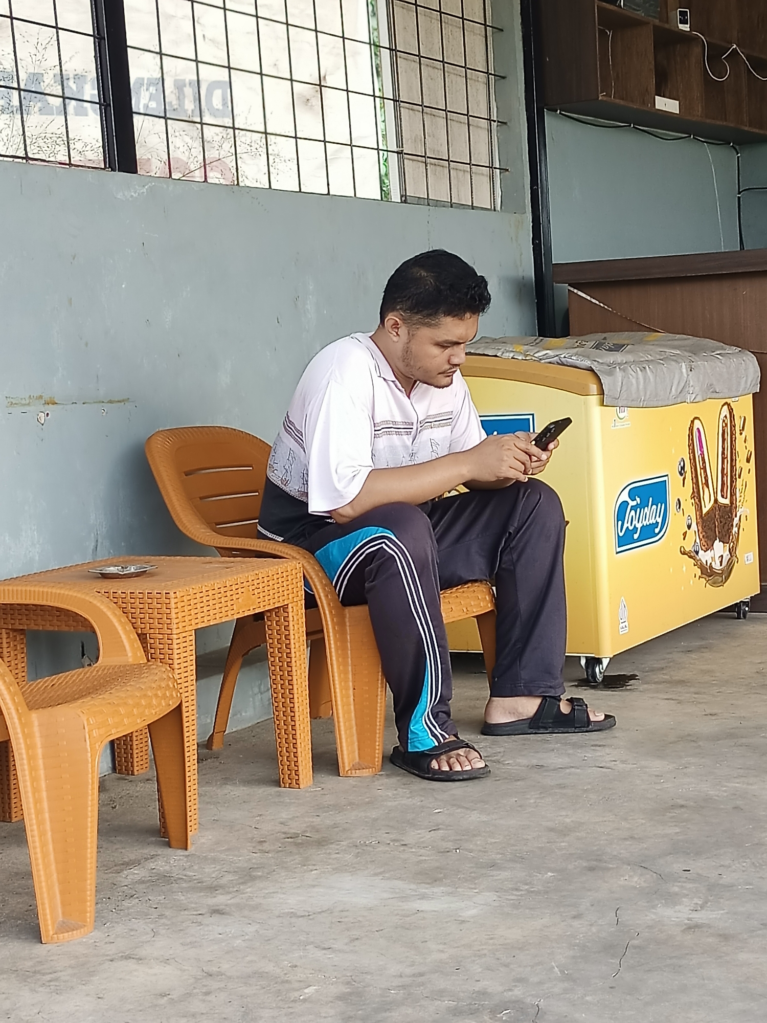 In Tajurhalang, Indonesia, a man sits on an orange plastic chair at a matching table, engrossed in his smartphone. He is wearing a white polo shirt with a striped collar and dark pants with a blue stripe down the side, and black sandals. To his right, a yellow ice cream freezer with a "Joyday" logo is visible, and further to the right, a wooden shelf unit is mounted on a teal wall. The scene takes place outdoors on a concrete floor, with a grey wall and a barred window in the background.