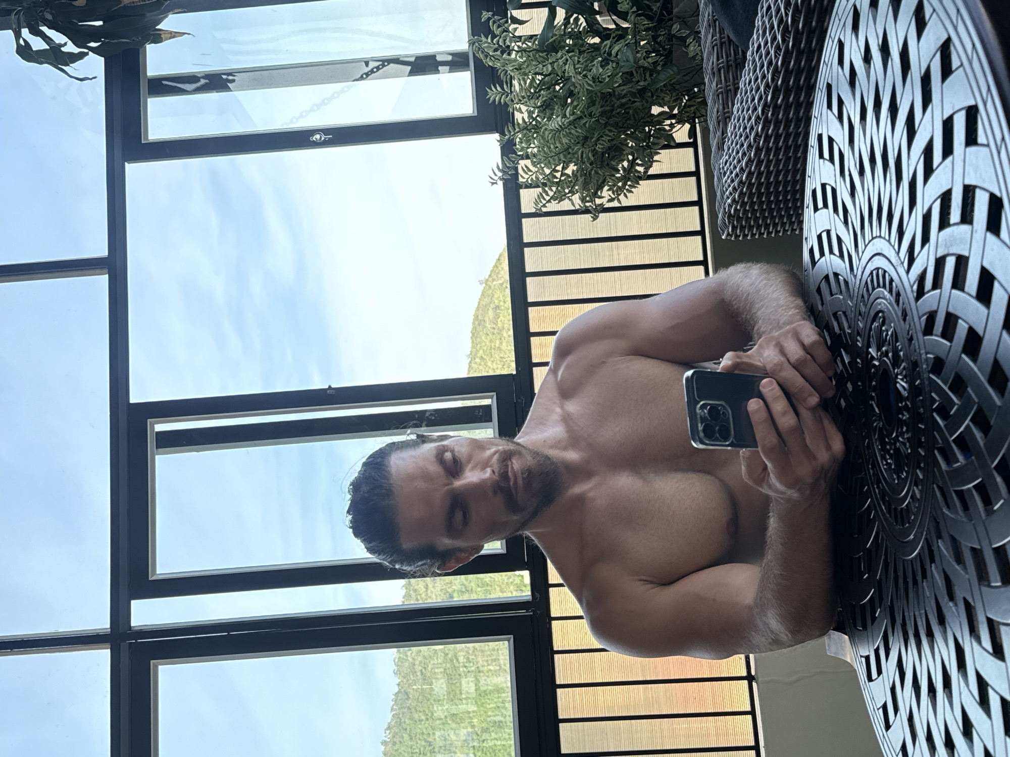 A shirtless man with a beard and hair tied back sits at a round, intricately designed table, looking down at his phone. He is bare-chested, with visible muscles. The table is made of dark metal. Behind him, a large window with black framing reveals a green, hilly landscape under a bright, cloudy sky. A chain is visible hanging from the window frame on the right. To the right of the man, a lush green plant sits next to a wicker chair. The scene appears to be on a balcony or in a room with a view. The location is identified as Bac Nha Trang Ward, Vietnam.
