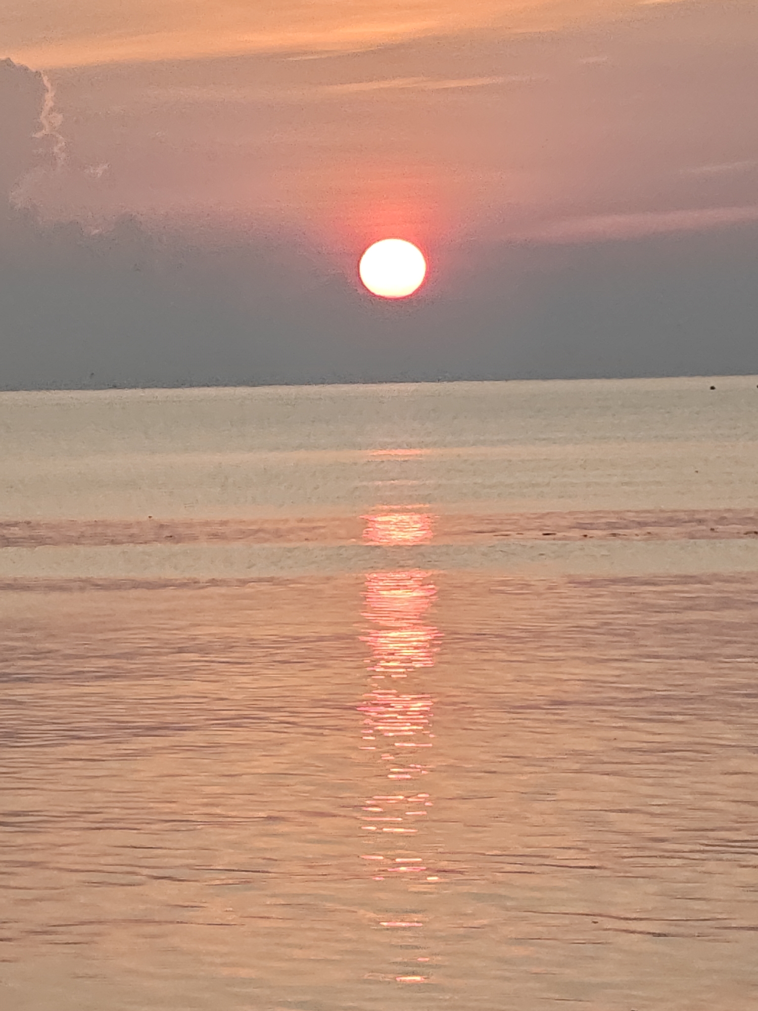 The image captures a sunset over the ocean. A pale pink sky transitions to gray towards the horizon, where the sun, a bright red-orange sphere, is positioned. A long reflection of the sun's light stretches down the surface of the water. The water is calm, with subtle ripples. The scene appears to be set in Lhokseumawe, Indonesia, due to the coastal setting and sunset. There are no people or specific objects.