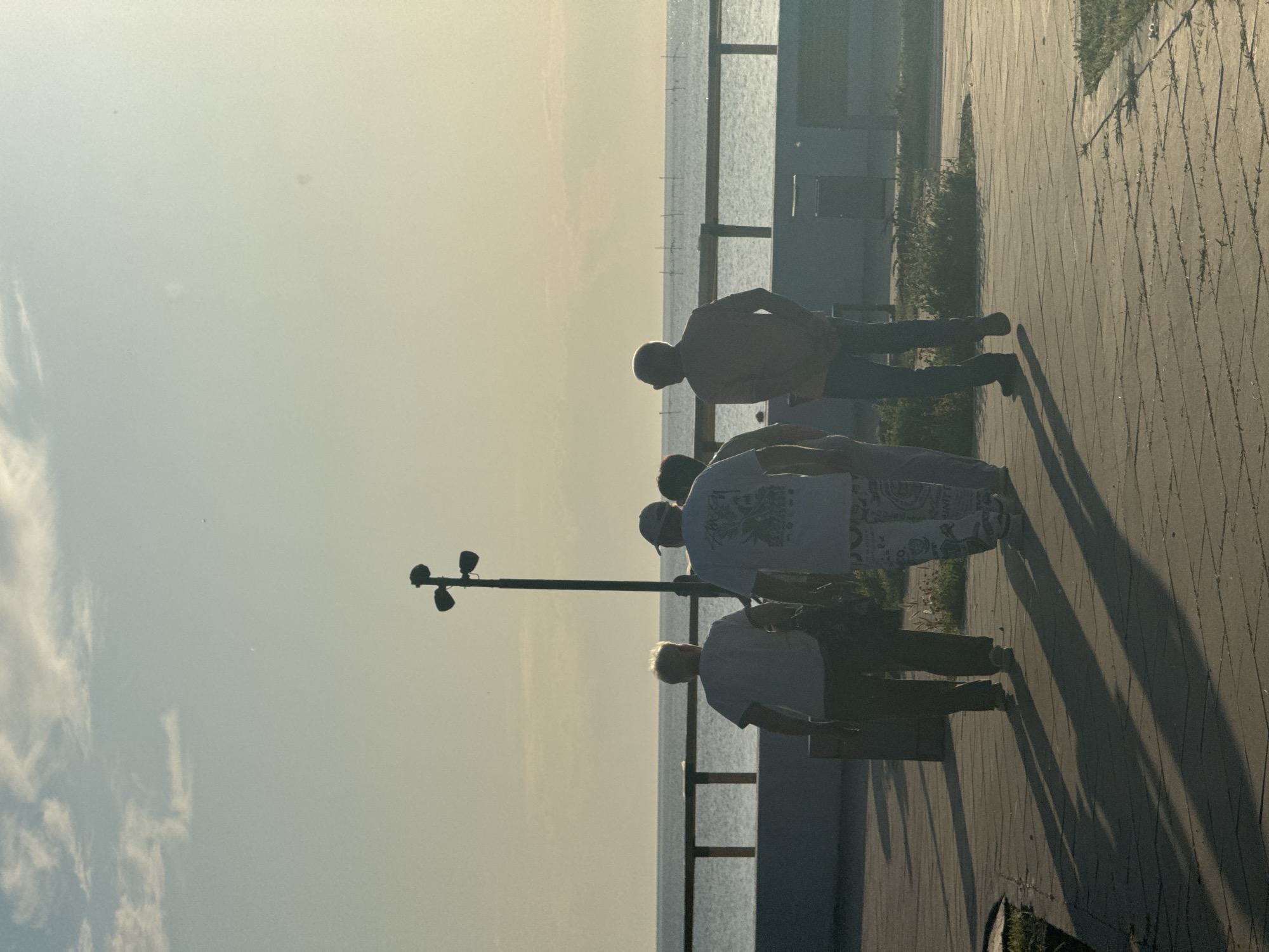 Four individuals are depicted walking away from the camera on a paved outdoor promenade in городское поселение Щелкино, Ukraine. The scene is illuminated by a low-angle sun, casting long shadows of the subjects towards the foreground. The sky above is mostly clear with a few wispy clouds.
From left to right, the first individual, an older adult with light-colored hair, wears a light t-shirt and dark pants, and appears to be carrying a dark object over their left shoulder. The second individual, an adult, wears a light-colored t-shirt with a dark graphic on the back, patterned light-colored pants, and a baseball cap worn backward. The third individual is partially obscured by the second and fourth individuals and wears a light-colored top. The fourth individual, an adult male, appears bald or closely shaven, wears an orange long-sleeved shirt, dark pants, and has hands in pockets. All four subjects are walking in the same direction, away from the viewer.
The foreground consists of a paved walkway made of rectangular blocks, with sparse vegetation growing between the cracks. A dark, multi-headed lamppost stands to the right of the first two individuals. In the midground, a dark railing system separates the promenade from a calm body of water, likely a sea or large lake, which extends to the horizon. Distant vertical structures, possibly poles for fishing nets, are visible on the water's surface towards the right. A light-colored low wall runs parallel to the railing, with some green foliage visible behind it. The overall lighting suggests late afternoon or early morning, with a warm, golden hue dominating the background sky and reflected on the water.