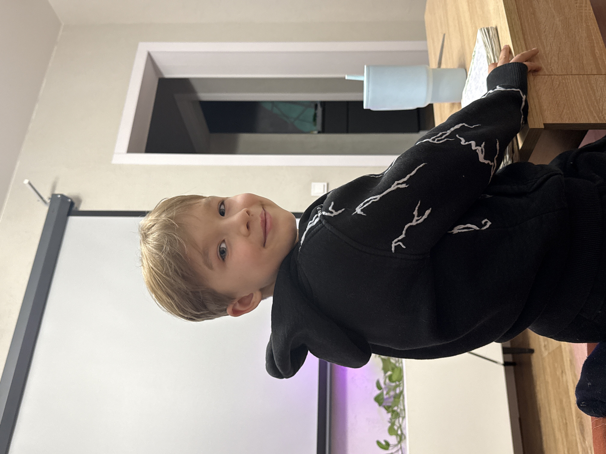 A young boy with light blonde hair and blue eyes is captured in a close-up, smiling directly at the camera. He appears to be a child, likely in his early elementary school years. He is wearing a black hooded sweatshirt adorned with white, lightning-bolt-like abstract patterns. His right arm is resting on a light-colored wooden table or desk, where a stack of papers or a book is visible beneath his hand.
To the right of his arm, a light blue insulated tumbler with a straw top sits on the wooden surface. The background reveals an indoor setting. On the left side of the frame, a large white projection screen or whiteboard is prominent, with part of a dark mounting bracket visible next to it. Below the screen, a soft purple glow illuminates some green leafy plants, possibly within an aquarium or a similar setup. High on the wall in the upper background, a recessed window or opening with a white frame is visible, showing a dark interior or exterior beyond, suggesting it might be evening or an artificially lit space. The boy's expression conveys happiness or contentment. No discernible text is visible in the image. The location context provided is Rostov-on-Don, Russia, although no specific visual cues within the image confirm this.