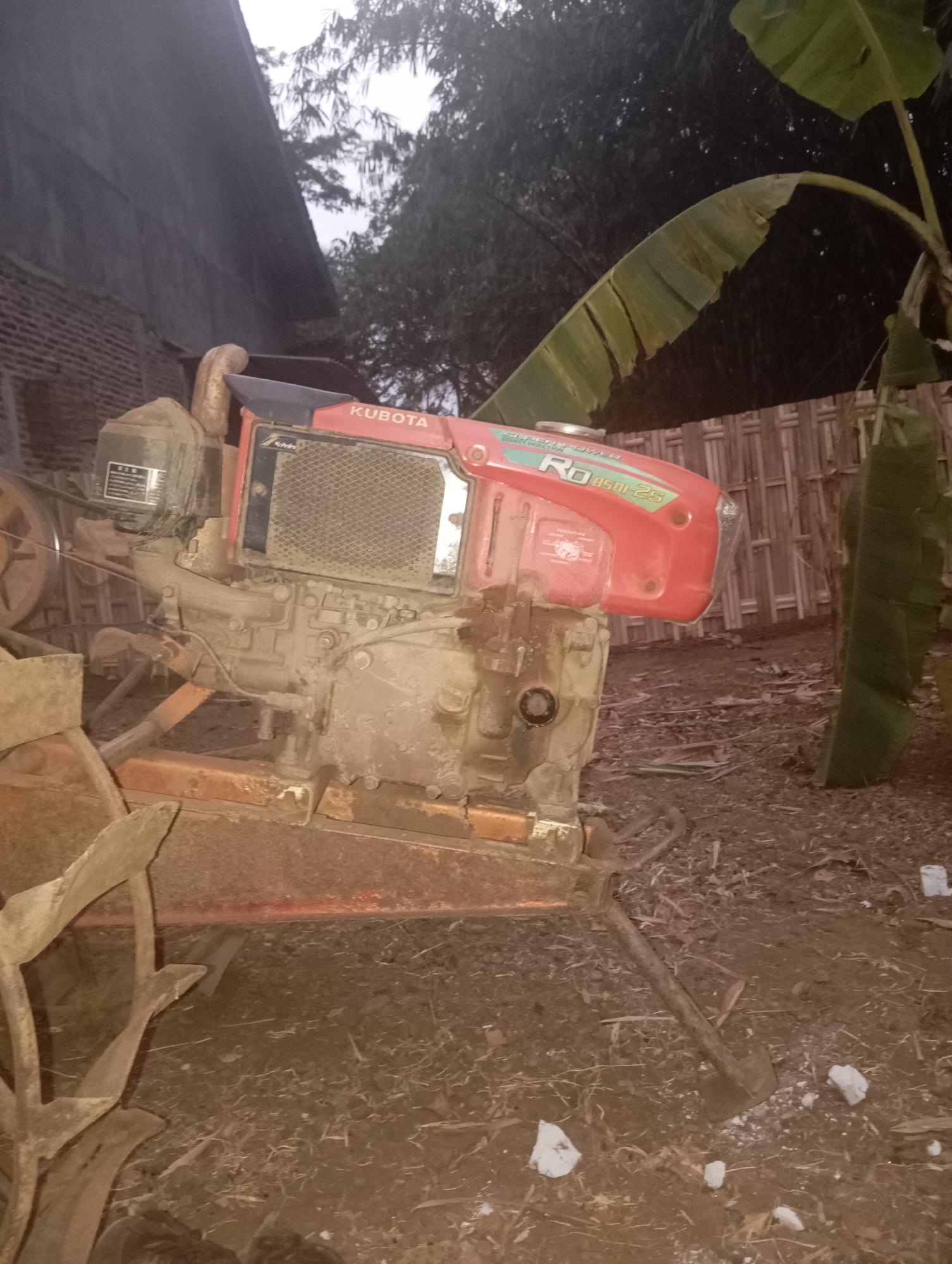 The image shows a Kubota RD8501-2S tiller parked on a dirt surface, likely in an agricultural or rural setting. The tiller is red and dirty, with a visible engine, fan, and tilling attachments. Behind the tiller, there is a brick wall and a thatched fence, with banana leaves and trees in the background. The scene appears to be outdoors, with natural lighting suggesting it might be late afternoon or early evening. There are no people visible in the image. The location is identified as Kedungbenda, Indonesia.