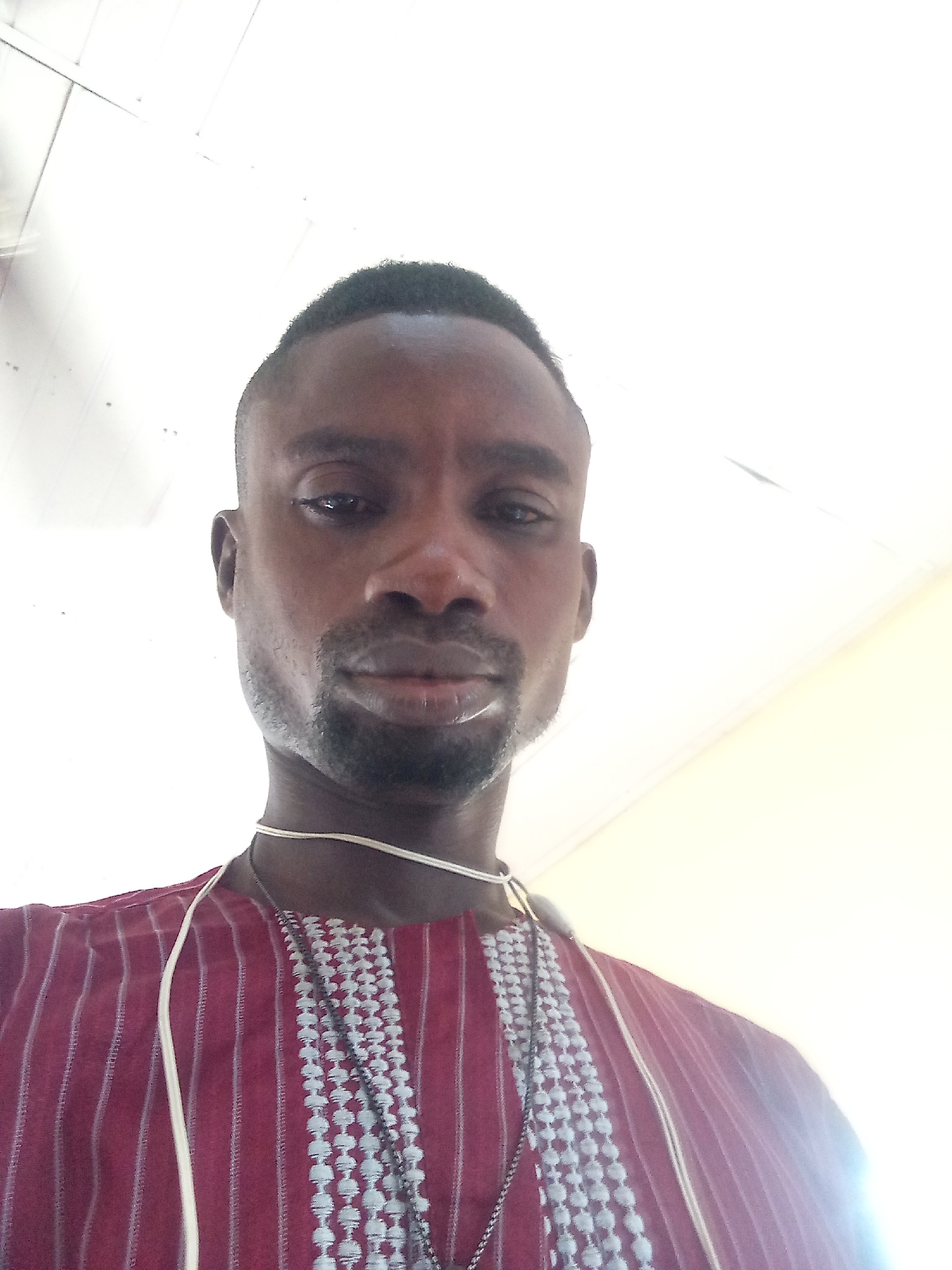 A selfie taken from a low angle shows a man wearing a dark red, striped garment with white and grey embroidery down the front. He has short, dark hair and a goatee. The man is looking directly at the camera. The background appears to be a white or light-colored ceiling with visible seams or panels. The image quality is somewhat blurry and overexposed, particularly in the upper portion. The man is wearing a thin, light-colored cord around his neck, with a layered necklace hanging over his garment. The scene is indoors.