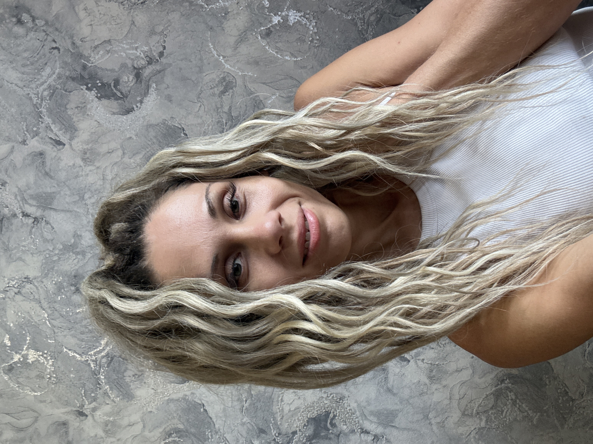 A woman with blonde, wavy hair pulled back from her face is looking at the camera. She is wearing a white tank top. She is positioned in front of a gray, textured wall. The image was taken in Rusanovo, Russia.