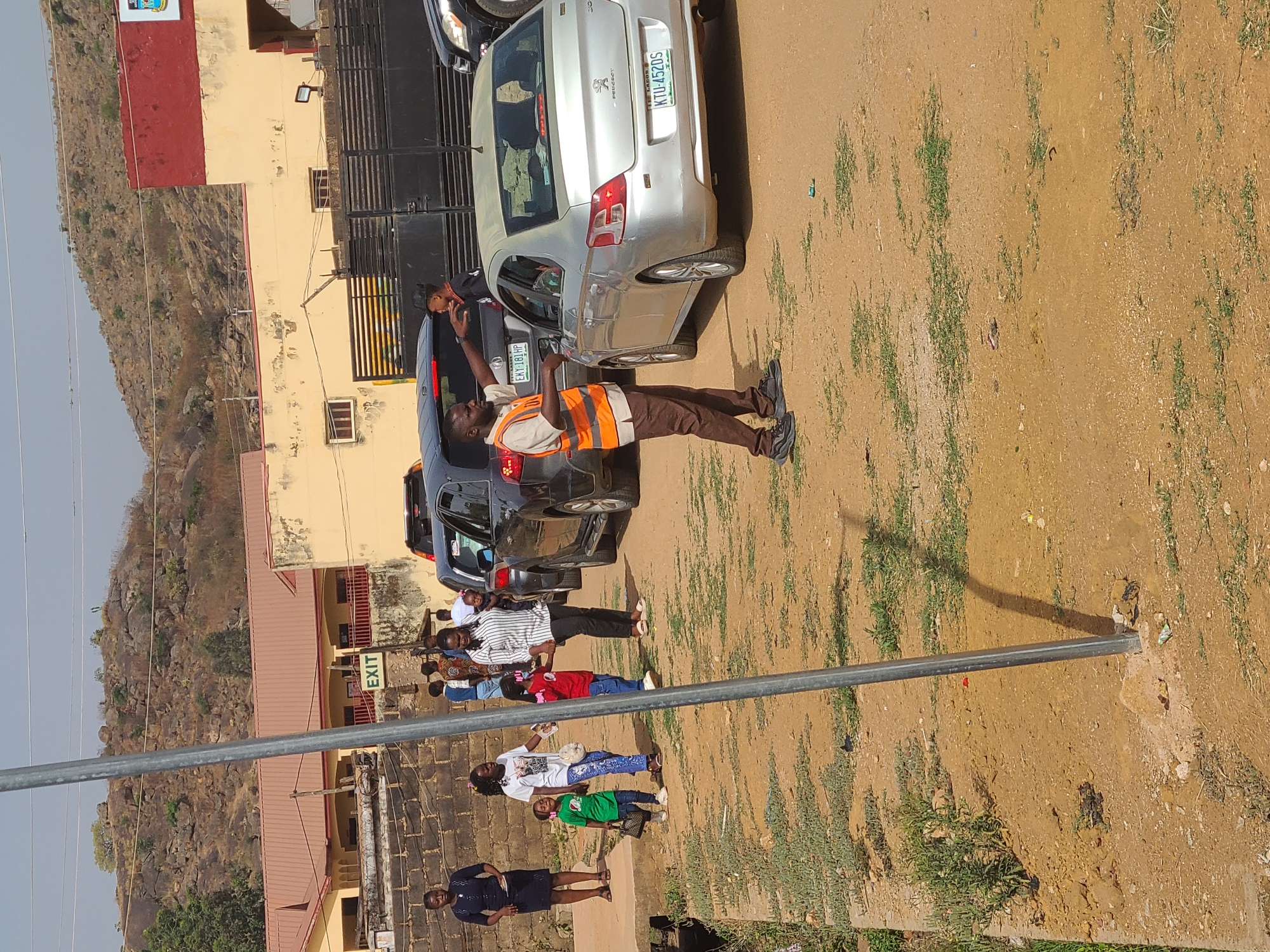 The image shows a street scene in Jos, Nigeria. A man wearing an orange safety vest is standing next to a silver Peugeot car, possibly directing traffic. Several other parked cars are visible behind the Peugeot. A group of people, including women and children, are standing or walking near a building with an "EXIT" sign. A rocky hillside is visible in the background. The ground appears to be a dirt surface. A long metal pole is in the foreground.