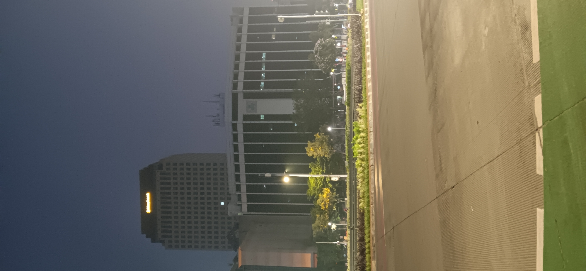 The image captures a dimly lit urban scene at dusk or night in South Jakarta, Indonesia. In the background, two prominent buildings stand out. The taller, left building features a glowing yellow sign at its apex that reads "Maybank". This building has a grid-like facade with numerous windows. To its right, a wider, more curved building is visible, characterized by vertical dark glass panels and a lighter, central section with a green emblem. Several antennas are mounted on the roof of this structure.
In the mid-ground, a well-maintained green space with trees and landscaping separates the road from the buildings. Streetlights illuminate the area, casting a soft glow on the vegetation and the pavement. The foreground is dominated by a multi-lane road, with visible lane markings and a textured surface, suggesting it is an asphalt road. A strip of green turf borders the road in the very bottom of the frame. The overall atmosphere is tranquil and slightly hazy, typical of an urban environment at night. There are no discernible people or vehicles in the scene.