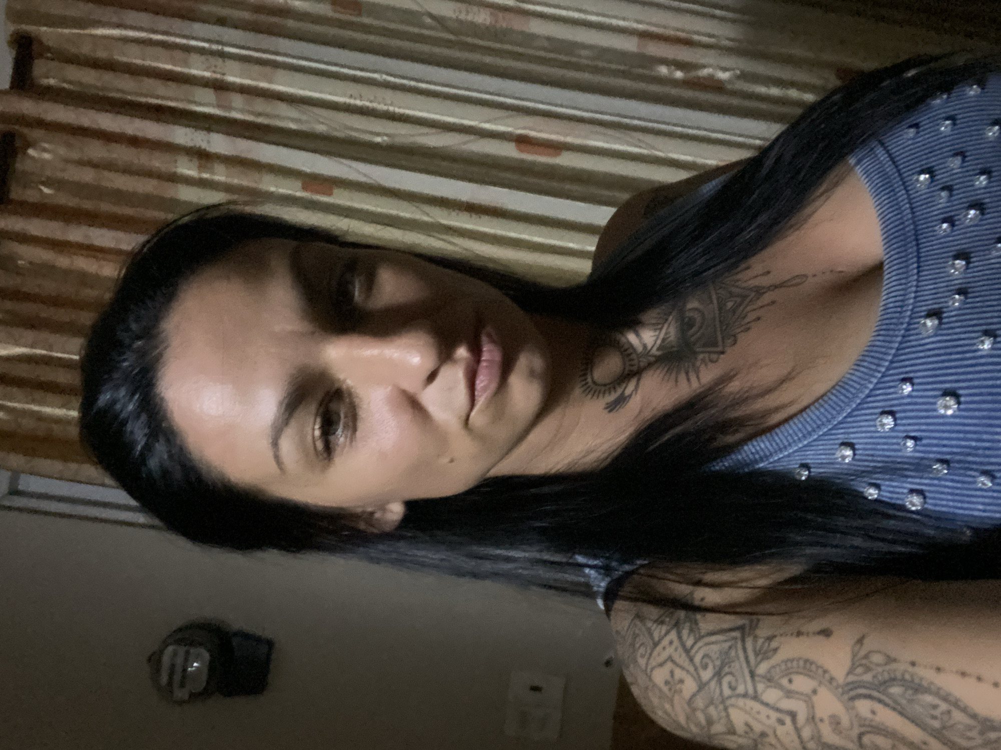 A woman with dark hair and tattoos is photographed indoors. She is wearing a blue shirt with sparkling embellishments and has a tattoo on her neck and another on her arm. The background includes a wall with a light switch and curtains. The location is Mũi Né, Vietnam.