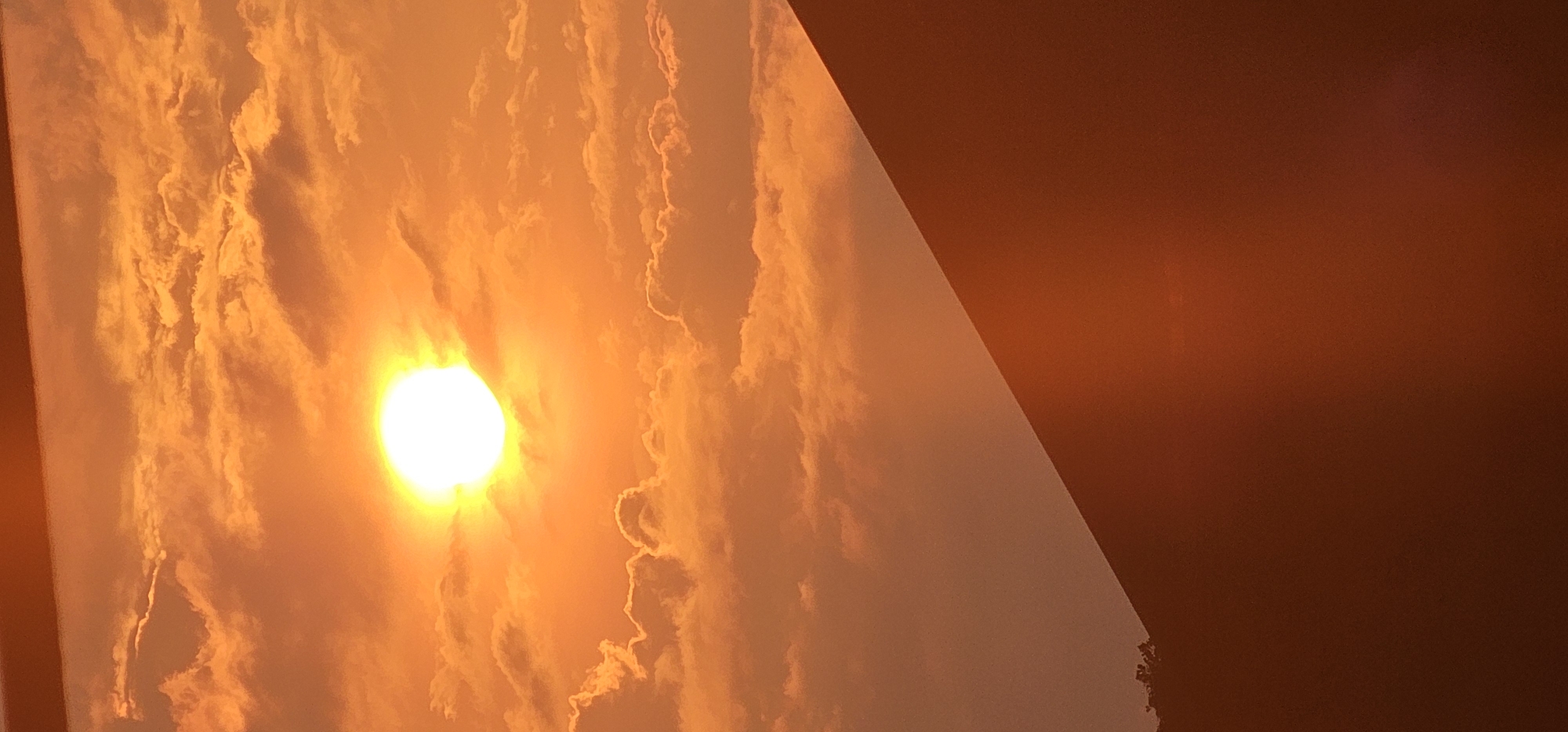 This is a photograph taken from a high vantage point, likely a mountain or elevated structure, looking towards a sunset. The sun, a bright, luminous orb, is prominently positioned in the upper center of the frame, casting a warm, orange glow across the sky. The sky is filled with scattered clouds, tinged with the same sunset hues, creating a dramatic and vibrant atmosphere. In the foreground, a dark, sloping surface dominates the lower portion of the image, suggesting the side of a mountain or a steep incline. A hint of foliage or trees can be seen on the far left edge of this surface, partially obscured by shadow. The overall impression is one of a serene and majestic natural landscape during a colorful sunset. The location is Puncak Jaya, Indonesia.