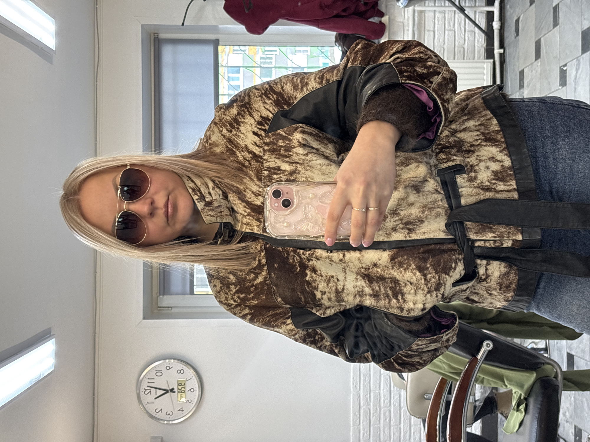 A woman is taking a selfie indoors. She has blonde hair and is wearing sunglasses and a patterned jacket. She is holding a phone in her right hand. In the background, there is a window, a clock on the wall showing the time as 8:58, a coat hanging, and some chairs. The location is Izhevsk, Russia.