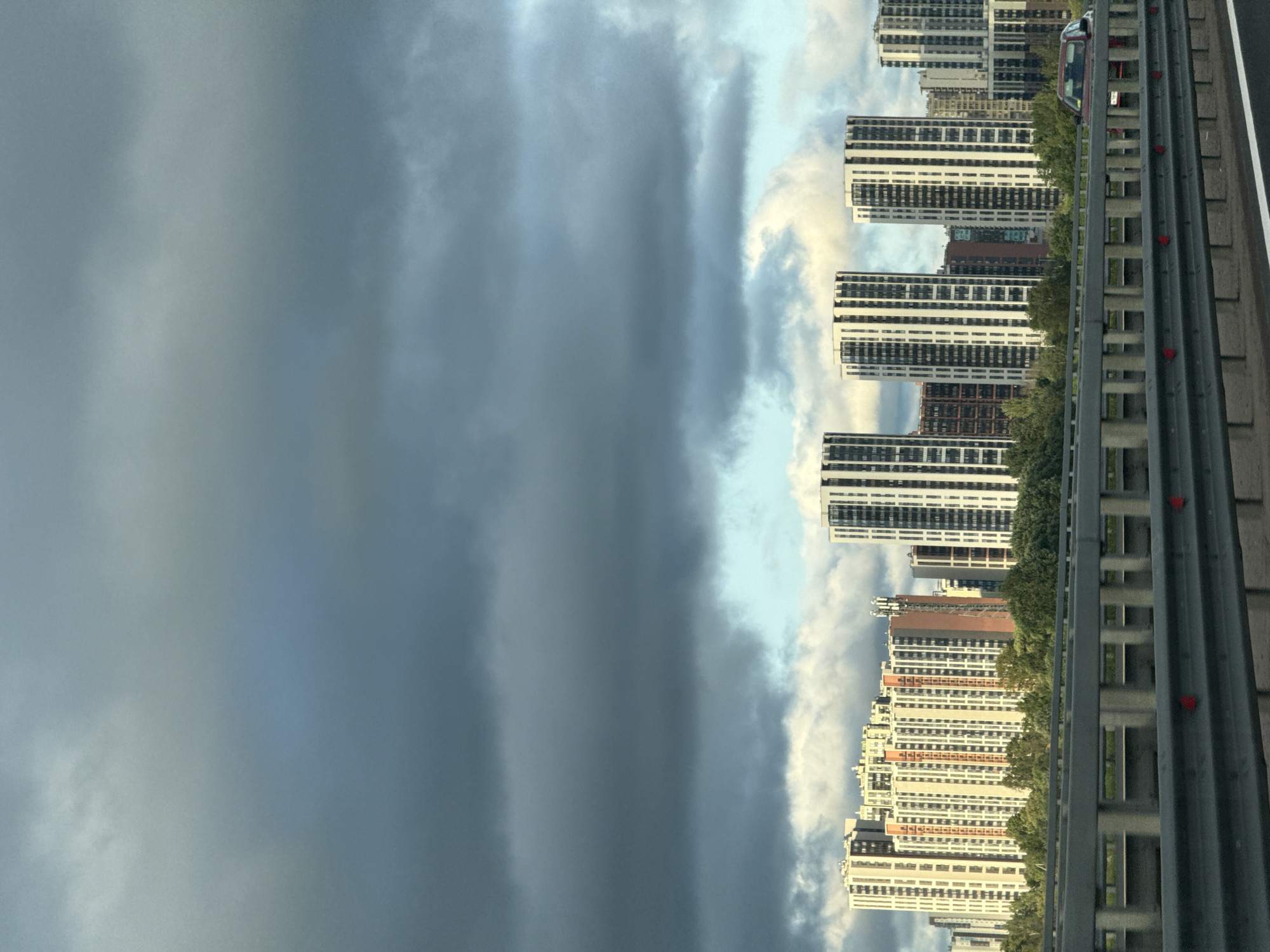 The image shows a modern cityscape with numerous high-rise buildings under a cloudy sky. In the foreground, a highway barrier is visible, with a red car driving on the road beyond it. The buildings are a mix of residential complexes, featuring balconies and window patterns, with some in lighter colors and others in darker shades. Trees line the area between the road and the buildings. The sky is dominated by dramatic, dark clouds with lighter patches peeking through, suggesting an overcast or approaching stormy weather. The overall scene conveys a sense of urban development and vastness.