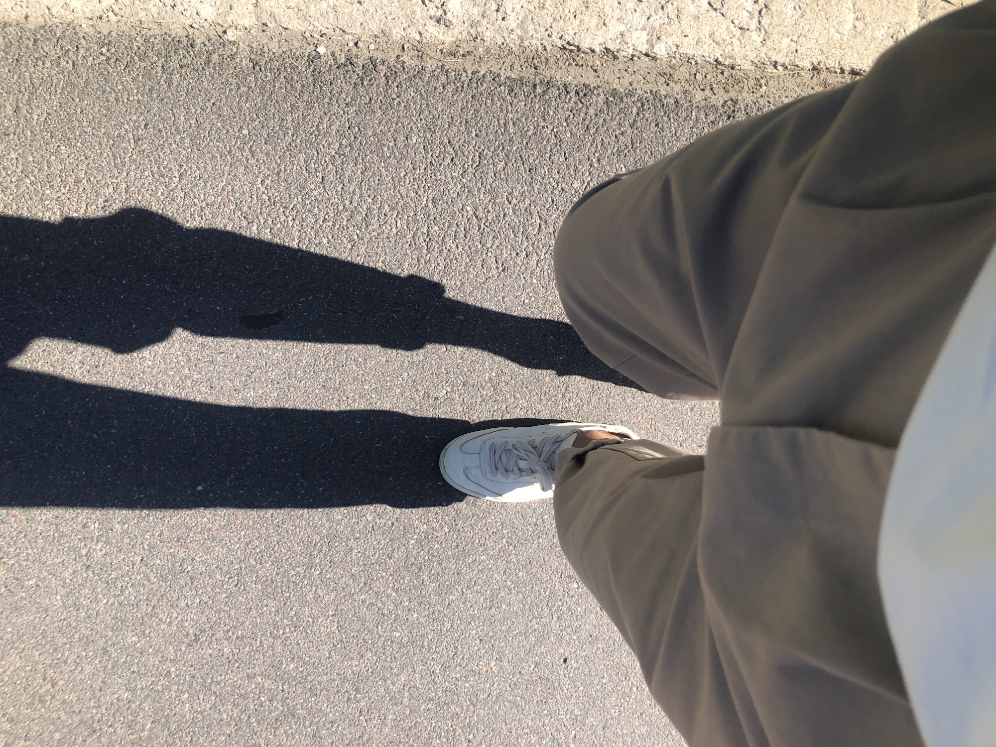 The image shows a first-person perspective of someone walking on an asphalt surface. The person is wearing light-colored sneakers and light-colored pants. The shadows of the person's legs are cast on the ground in front of them. The environment appears to be outdoors, possibly a sidewalk or road, with a sandy or gravelly edge on the right side. There is no specific visual information in the image to indicate a location in Saint Petersburg, Russia.