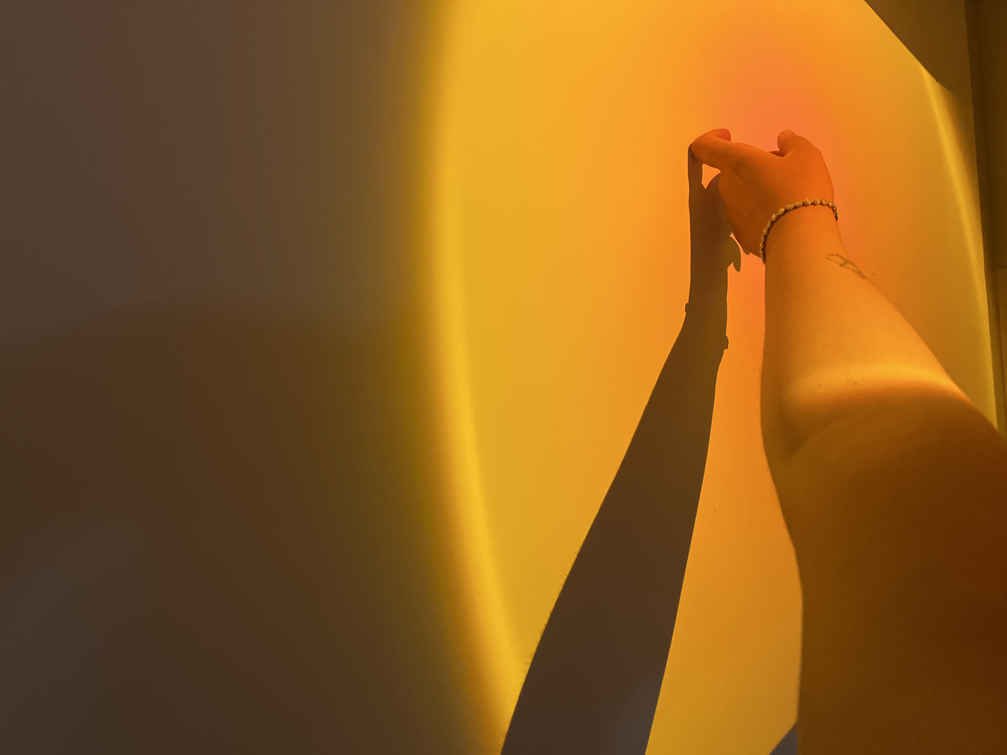 The image captures an indoor scene in Kiryat Bialik, Israel, illuminated by a strong, artificial warm light source projecting an orange-yellow gradient onto a vertical surface. A distinct bright yellow arc of light is visible across the illuminated area. A human right arm and hand are centrally positioned in the lower frame. The hand, wearing a bracelet on the wrist and displaying a faint tattoo on the forearm, is holding a small, light-colored, translucent object between its thumb and forefinger. A prominent, dark shadow of an arm and hand is cast onto the illuminated surface, originating from the left side of the frame. The upper portion of the background is a darker brown, transitioning to the vibrant orange-yellow illumination below.