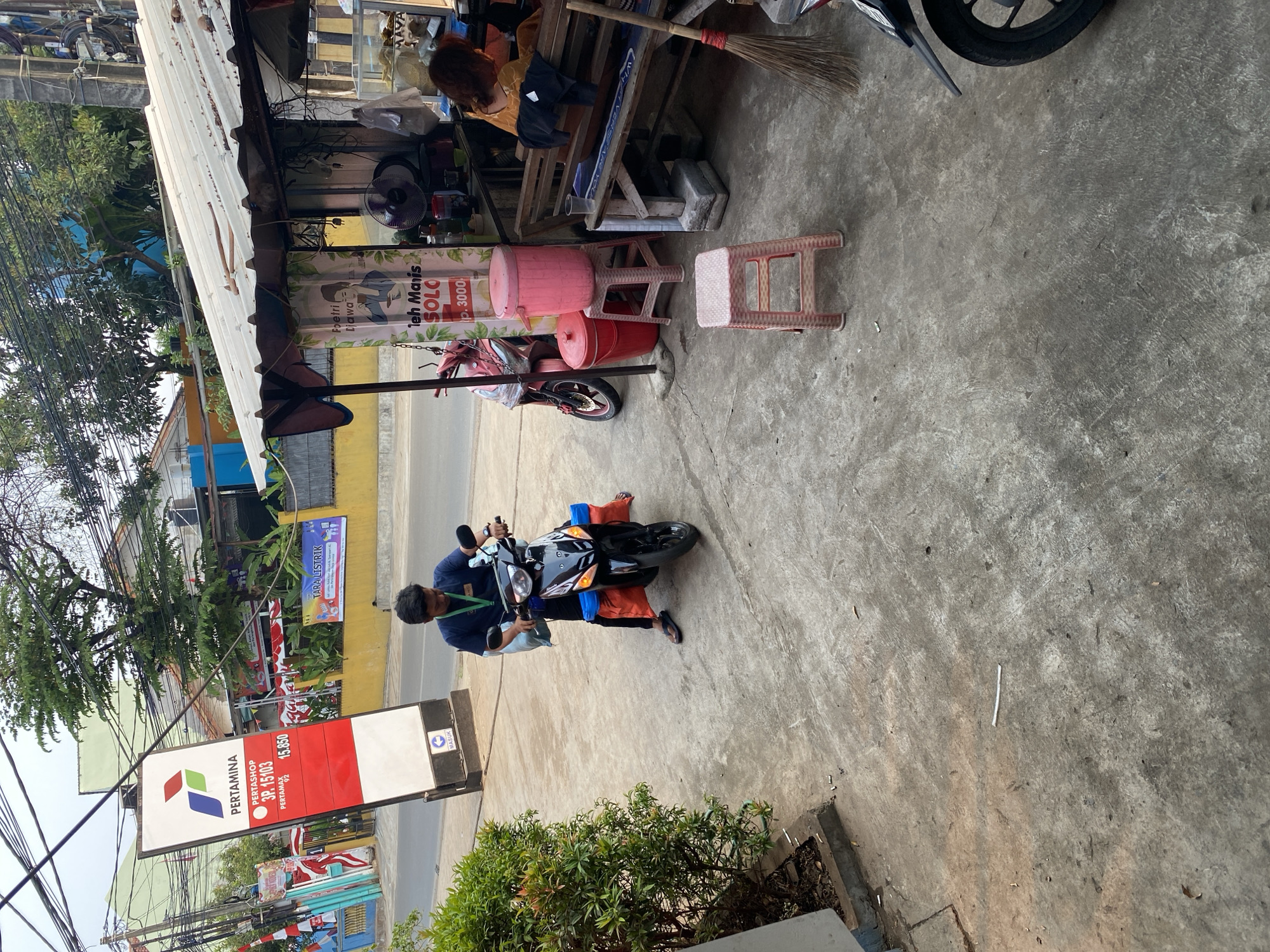 The image depicts a street scene in Tangerang, Indonesia, likely near a Pertamina gas station. In the foreground, a young man wearing a dark blue shirt and black pants is seated on a black motorcycle. He is holding a blue bag and has orange bags attached to the rear of the motorcycle. The motorcycle is parked on a concrete sidewalk.
To the left of the man and motorcycle, a tall, red and white Pertamina sign stands. The sign displays the Pertamina logo and information about "Pertamax" fuel prices. Adjacent to the sign is a lush green bush.
Behind the man and motorcycle, there is a small roadside stall with a white corrugated metal roof. The stall appears to sell drinks, with a sign featuring an illustration of a woman and text that includes "leh Manis SOLO" and "Rp. 3000". Red and pink plastic buckets and stools are placed outside the stall. A pink trash can is on top of a stool, and a red bucket is on the ground.
A woman with reddish-brown hair, wearing a yellow shirt, is visible seated at the stall, interacting with someone inside. A broom leans against the stall. Further in the background, there are buildings, trees, and overhead electrical wires, suggesting an urban environment. The ground is paved with concrete. The overall scene suggests a typical day at a local convenience stall or eatery alongside a busy street.