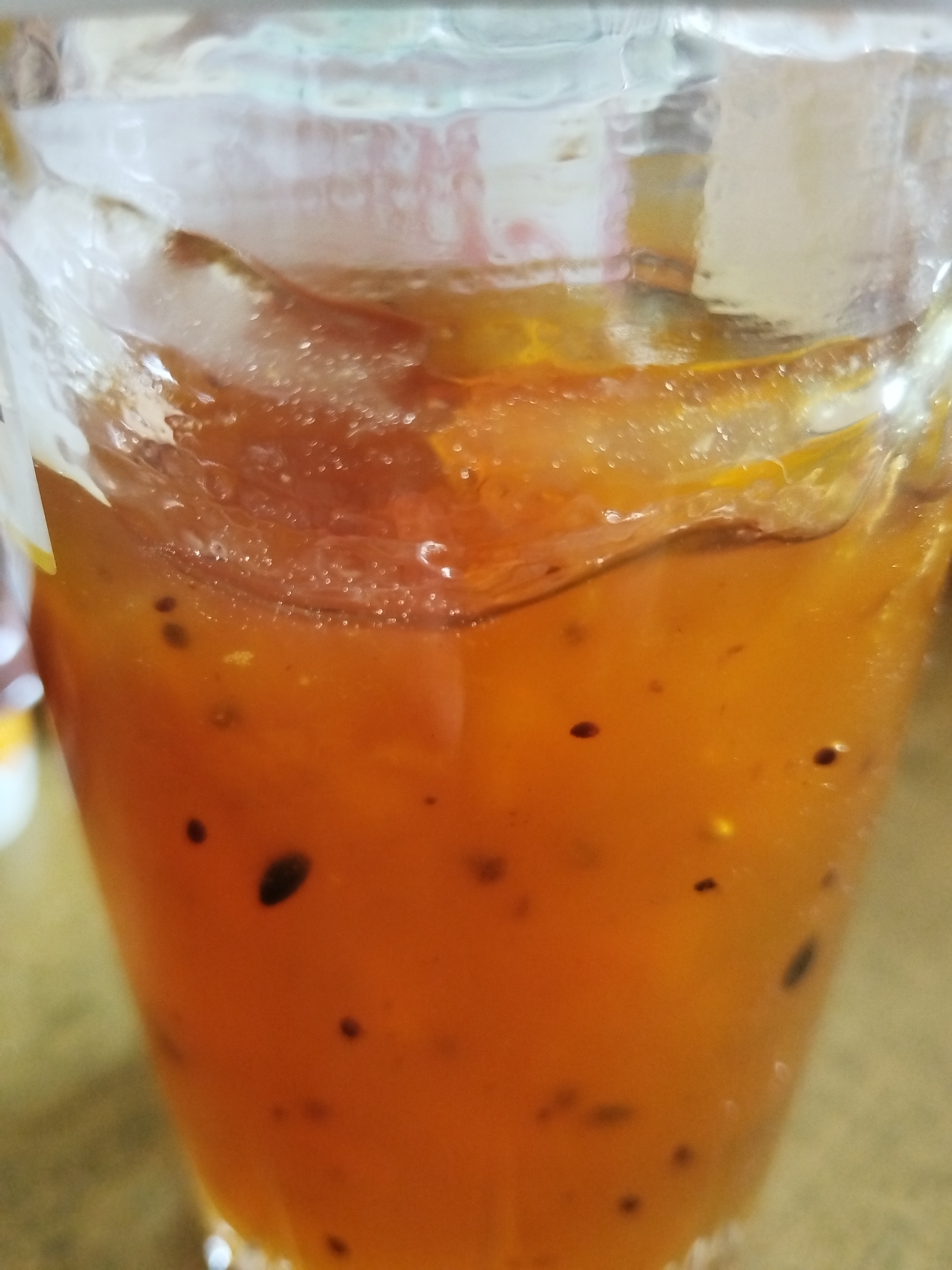 A close-up shot shows a glass filled with an orange liquid containing dark seeds. The liquid appears to have ice in it. The glass is transparent, and condensation is visible on its exterior. The background is slightly blurred. The location is unknown.