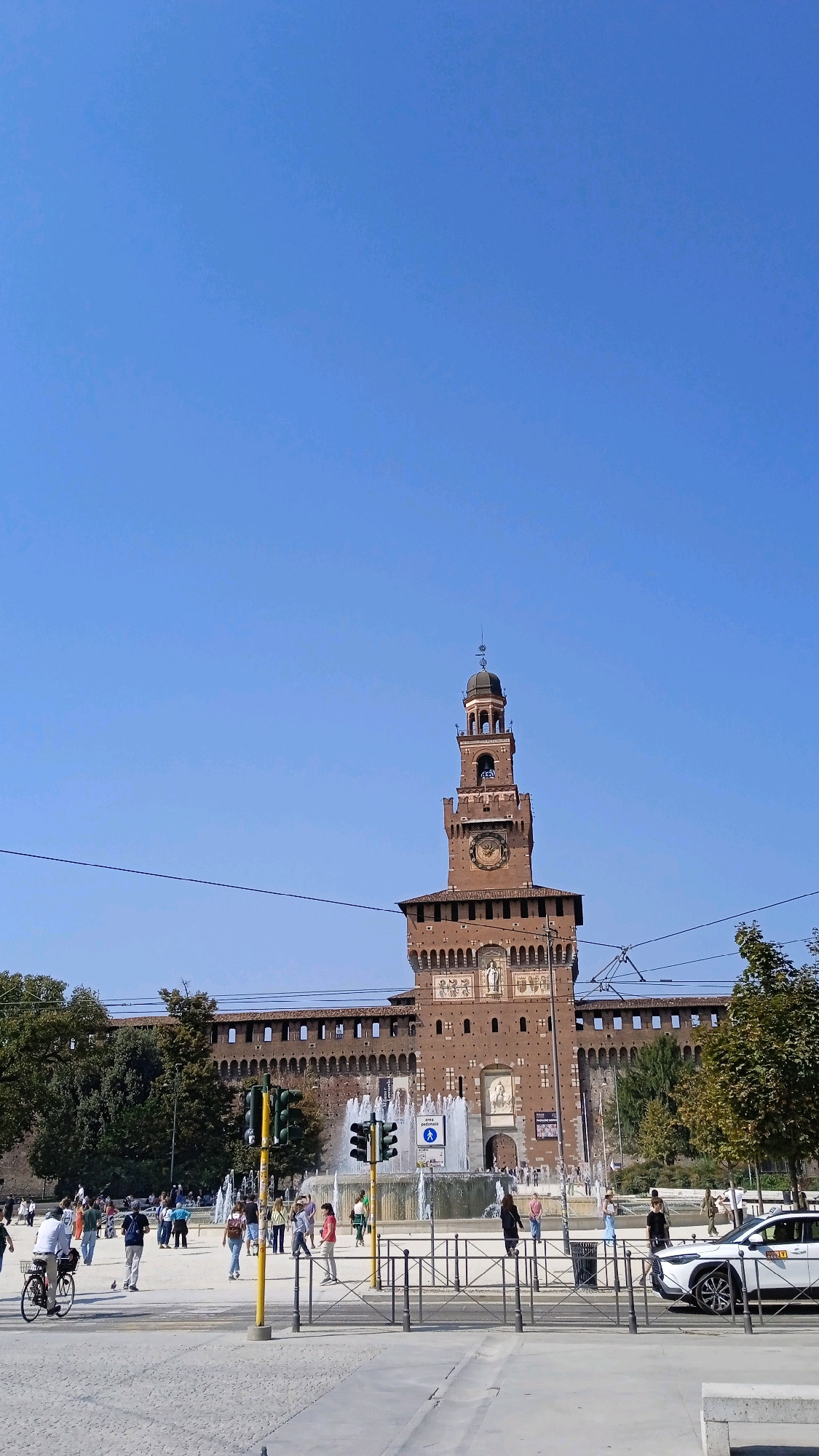 The image captures a sunny day at Sforza Castle in Milan, Italy. The prominent brick tower of the castle, topped with a dome, dominates the upper center of the frame, featuring a large clock face. In front of the castle, a wide plaza is bustling with activity. A large fountain sprays water, with people visible walking around it. Several traffic lights and street lamps dot the scene, indicating a public thoroughfare. Pedestrians are seen in various locations within the plaza, some walking individually and others in small groups. A white car, possibly a taxi, is parked on the right side of the image. Lush green trees frame the castle walls on either side. The sky is a clear, vibrant blue.