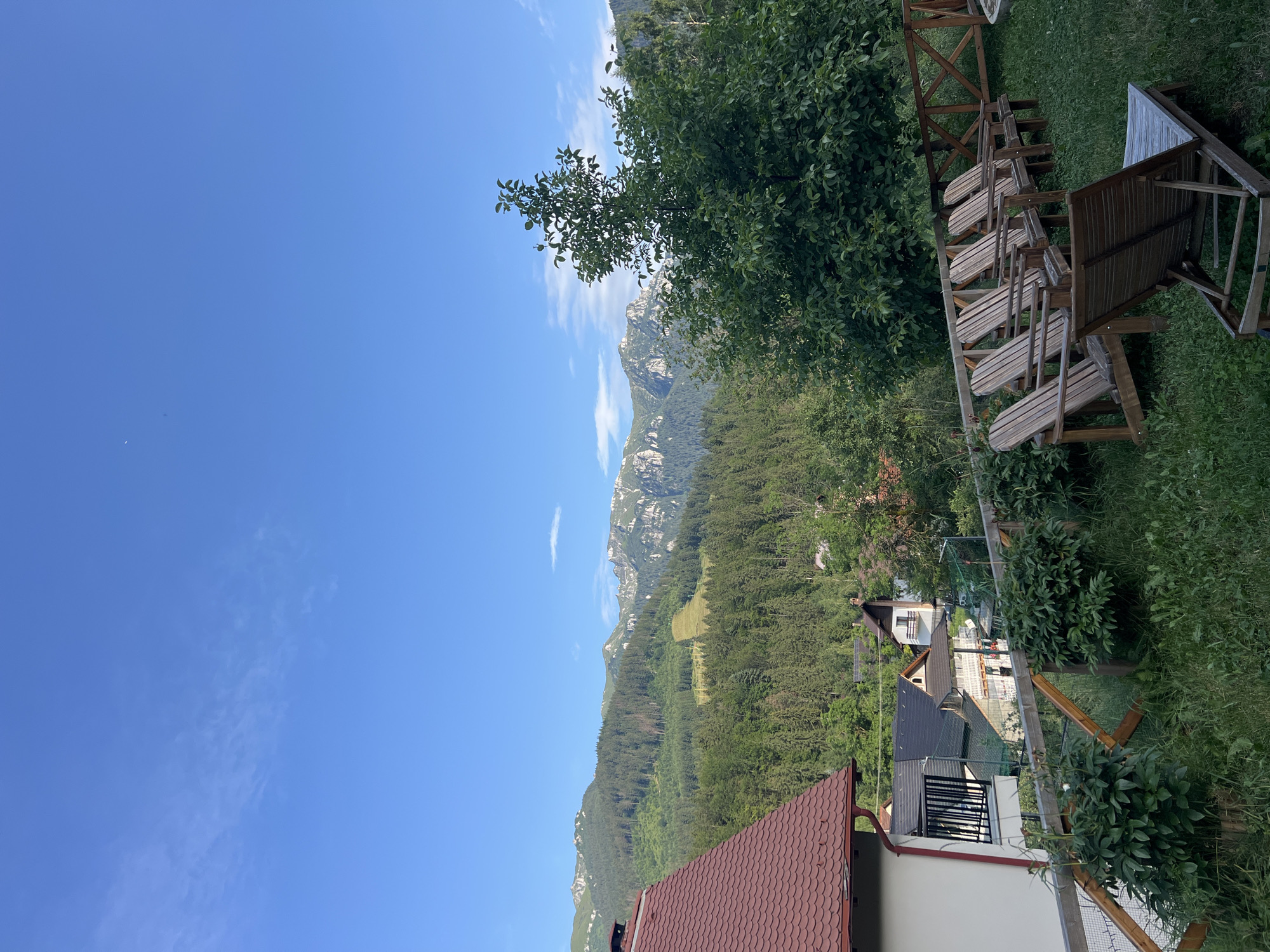 This media file shows a scenic mountain landscape with a clear blue sky and some scattered clouds. In the foreground, there is a wooden deck with several empty chairs, suggesting a place for relaxation or observation. Behind the deck, there is lush green vegetation and trees. Further in the distance, a dense forest covers the slopes of the mountains, which are rugged and rocky. Several houses with red tile roofs are visible nestled amongst the trees, indicating a human settlement in the mountainous region. The scene appears to be set during the daytime, likely a sunny day, given the bright sky and shadows. There is no text visible in the image. The overall impression is one of a tranquil and picturesque natural setting.