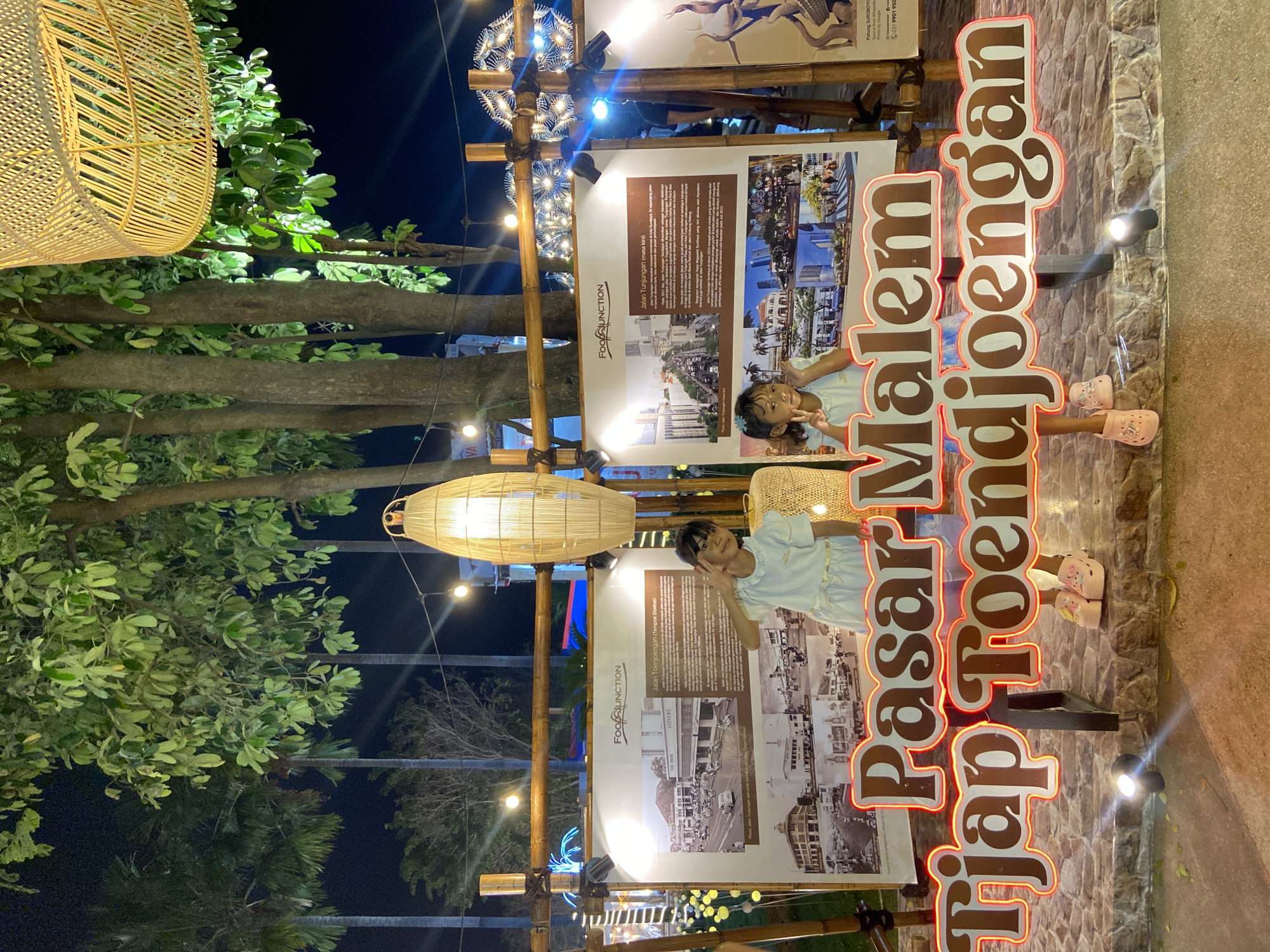 Here is a detailed description of the image:
The image appears to be taken at night in Surabaya, Indonesia. There are two young girls standing near a sign that reads "Pasar Malem Tjap Toendjoengan". The sign is illuminated with lights. Behind the girls are large posters or informational displays, one featuring historical images labeled "Jalan Toenjoengan (Tempoe Doeloe)" and another depicting modern views called "Jalan Tunjungan (masa kini)". The displays reference FOODJUNCTION and HOENKWE.
The location is decorated with bamboo structures and hanging woven lanterns. The environment includes trees and greenery, suggesting an outdoor or semi-outdoor setting. The overall scene seems festive and related to a market event (Pasar Malem).