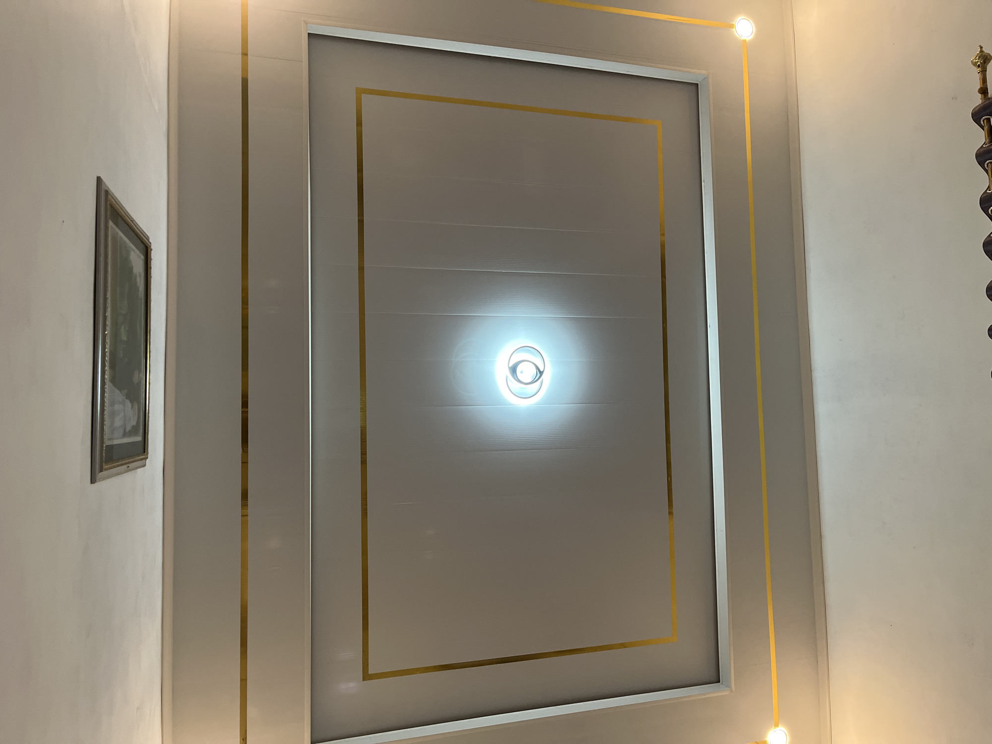This is a forensic, matter-of-fact description of a ceiling in Kebumen, Indonesia. The ceiling is made of white panels with gold trim. There is a rectangular recessed area in the ceiling with a framed picture. A light fixture is visible in the center of the recessed area. Two small spotlights are positioned along the lower edge of the ceiling, one on the left and one on the right. The lower portion of the image shows a white wall with a curtain rod and dark curtains.