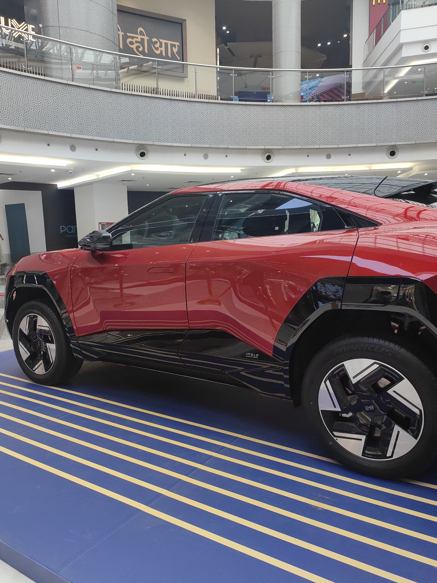 A red electric vehicle is displayed on a blue platform with yellow stripes in a shopping mall. The vehicle has black accents on its lower body and wheel arches. The front wheel is visible on the left, and the rear wheel is visible on the right. The car's design features a sleek, modern aesthetic. In the background, there are levels of the mall with railings, escalators, and signage. One sign in Hindi appears to say "कही आर". The scene suggests a promotional event or showroom display. The location is the Phoenix Palladium mall in Mumbai, India.