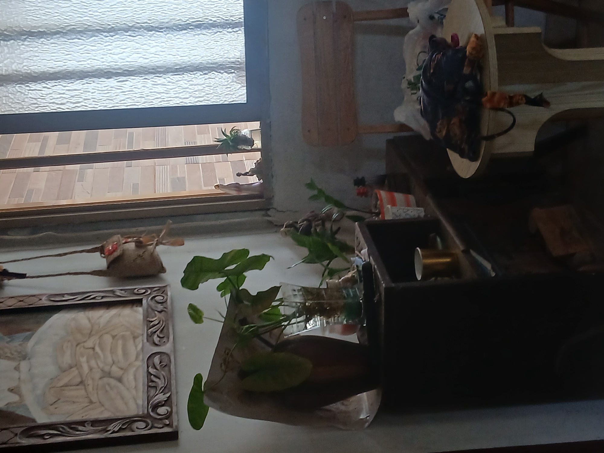 The image depicts an interior scene, likely a room in Santa Ana, Venezuela, characterized by a mix of decorative elements and household items. In the upper left, a framed artwork, possibly a bas-relief or carving, is visible, featuring figures with a beige tone. To its right, a small, rustic bag hangs by a string.
The central area of the image is dominated by a dark wooden cabinet or shelf unit. On top of this unit, several potted plants are arranged. One plant in a clear glass vase with water and pebbles stands prominently. Another plant is in a striped pot, and a third plant is seen in the background. Various other objects, including a metallic cup, are present on the shelves.
To the right of the cabinet, a wooden chair with a slatted back is partially visible. A round side table stands in the foreground, on which a pile of dark fabric items, possibly clothing or bags, is placed.
The background includes a window with a textured, translucent curtain, and the exterior wall of a building with a tiled pattern is visible through the window. A small figurine of a person is placed on the windowsill. The overall lighting suggests a dimly lit interior.