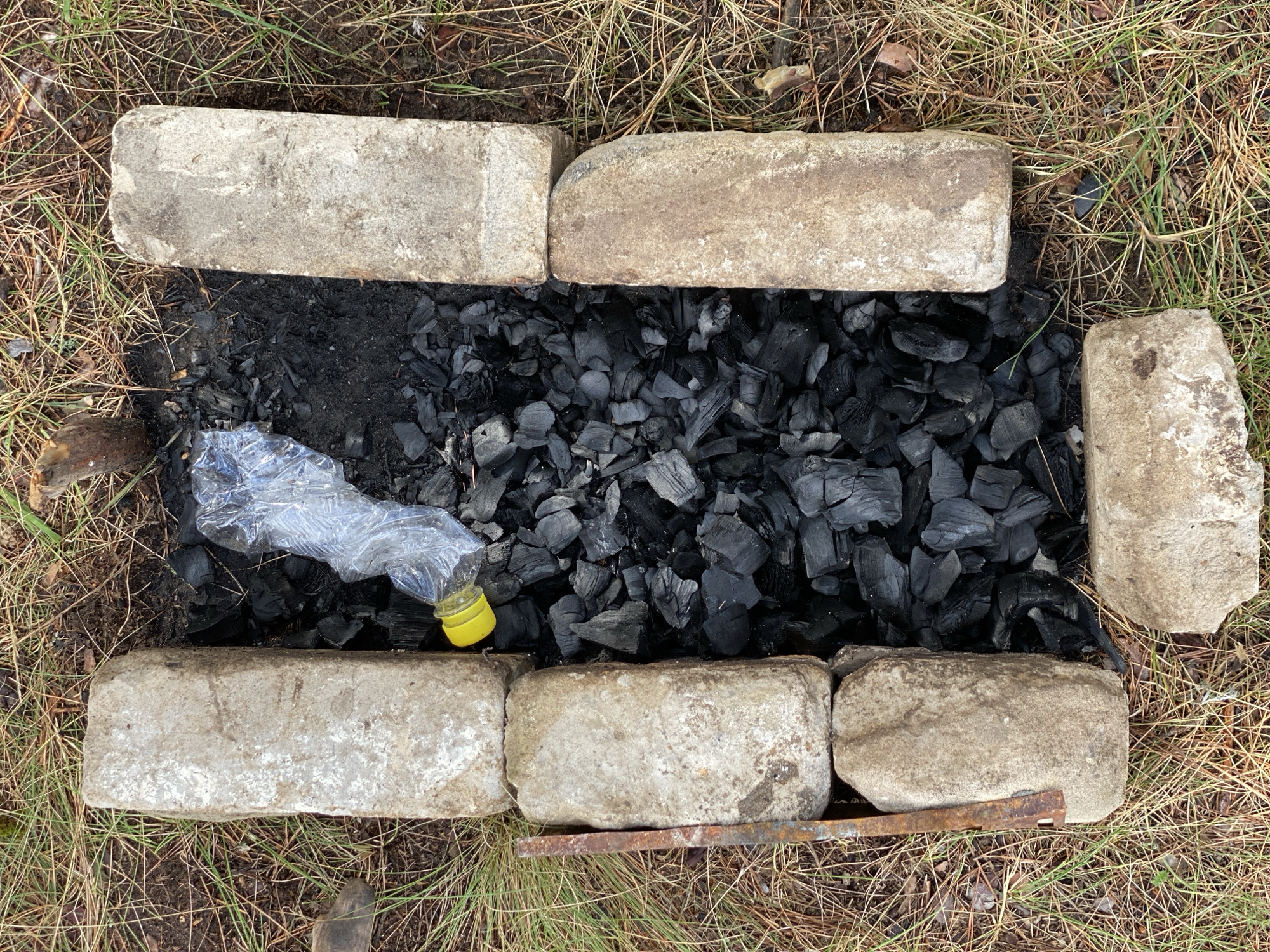An overhead view shows a makeshift fire pit constructed from concrete blocks, filled with charcoal. A crumpled plastic bottle with a yellow cap lies on top of the charcoal. The fire pit is surrounded by dry grass and pine needles, indicative of an outdoor, possibly wooded, environment. There are no people or any overt actions taking place within the frame. The scene appears to be in Leninsky, Russia, based on the provided location.