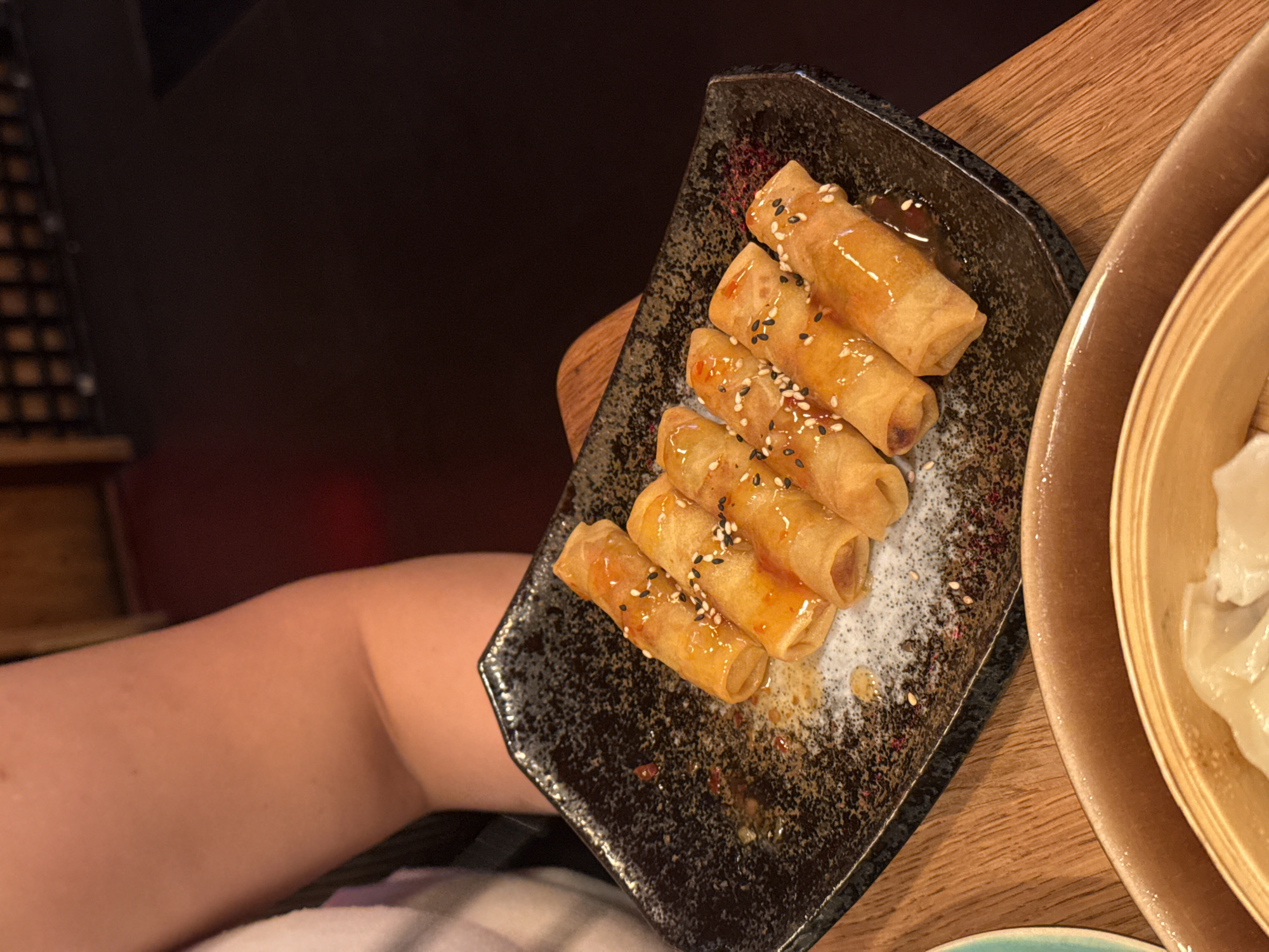 A slightly high-angle close-up photograph shows a black rectangular plate containing six small, golden-brown spring rolls, drizzled with a glossy sauce and garnished with black and white sesame seeds. The plate sits on a light-brown wooden table, partially overlapping a circular bamboo steamer containing dumplings. A person's arm and shoulder are visible in the lower left of the image, wearing what appears to be a white or light-colored garment. The background is blurry and dark, suggesting an indoor dining environment. The setting appears to be a restaurant, likely in Sofia, Bulgaria, based on the food served, but this is not explicitly discernible from the image itself.