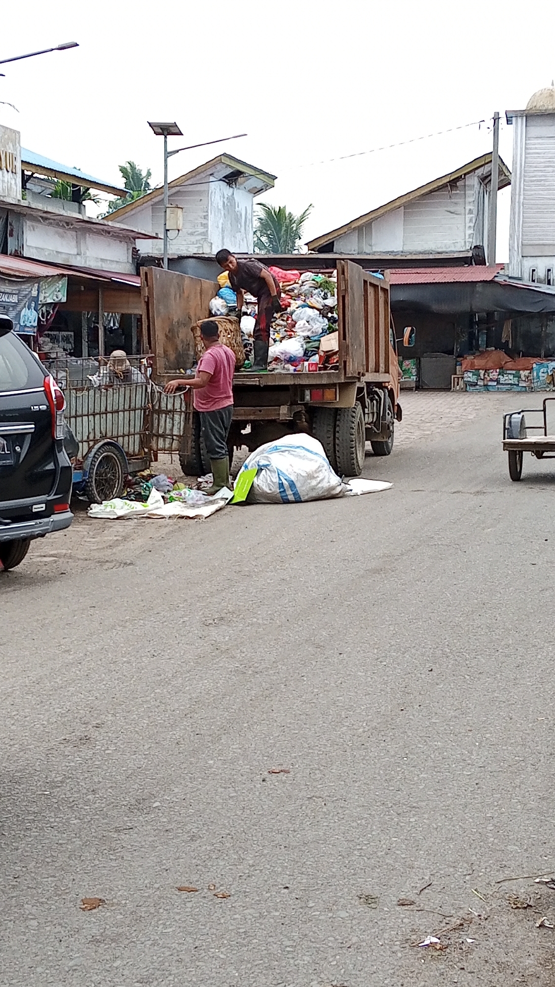 In Puloara, Indonesia, a scene unfolds on a paved street, suggesting an area of urban activity. Two individuals are engaged in waste management. One person, wearing a dark t-shirt and dark pants with green rubber boots, is positioned inside a large, rusty truck filled with an extensive amount of refuse. They appear to be actively sorting or loading the trash. The truck itself is substantial, with visible tires and a weathered appearance.
Beside the truck, another individual, dressed in a pink shirt and dark pants, also wearing green rubber boots, stands on the street. They are near a metal cart with a cage-like structure, also containing waste, and a separate large white and blue plastic bag of trash. Several other smaller pieces of trash and bags are scattered on the ground around the truck and cart, indicating the ongoing process of waste collection and disposal.
In the background, buildings with corrugated metal roofs and concrete walls are visible, suggesting a commercial or residential area. A black SUV is partially visible on the left side of the frame, adding to the sense of an active street scene. The sky is overcast. The overall impression is one of daily work related to waste management in a developing urban environment.