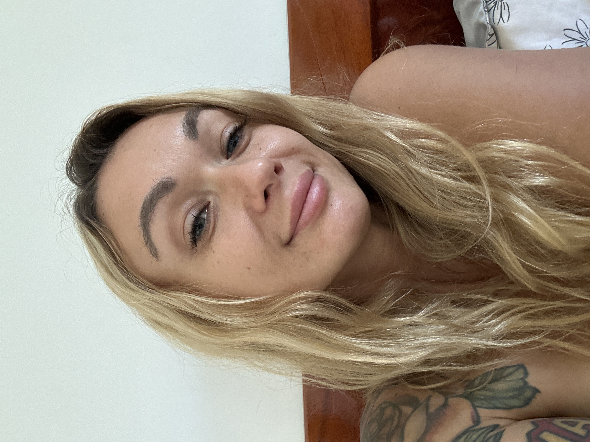 A close-up selfie shows a light-skinned woman with blonde, wavy hair. She is lying down, with the right side of her face angled towards the camera. Her features include well-defined eyebrows, light-colored eyes, and full lips. Freckles are visible on her face. A tattoo is partially visible on her left arm. The background includes a wooden headboard and a white pillow with a floral pattern. The setting appears to be indoors, likely a bedroom. There is no information to determine if the location is in Mui Ne, Vietnam.