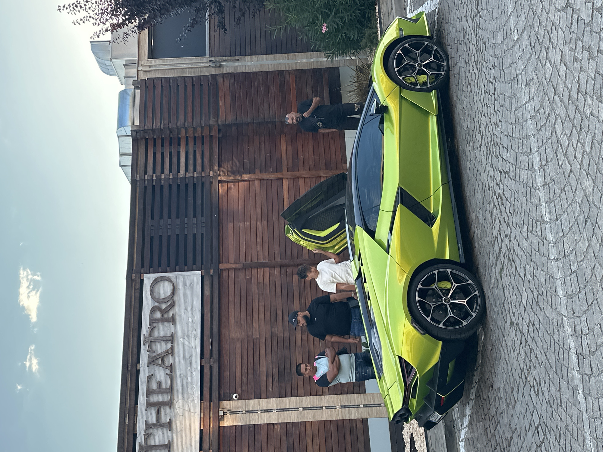A bright green Lamborghini Aventador is parked on a cobblestone street in front of a building with a wooden facade. The building has a sign that reads "TEATRO Piano Live." Four men are standing near the car. Three men are grouped to the left of the car, and one man is standing to the right, talking on his phone. The Lamborghini's doors are open, revealing its interior and the lime green accents on the doors and seats. The sky is a pale blue with a few scattered clouds. The overall scene suggests a luxurious and social setting.