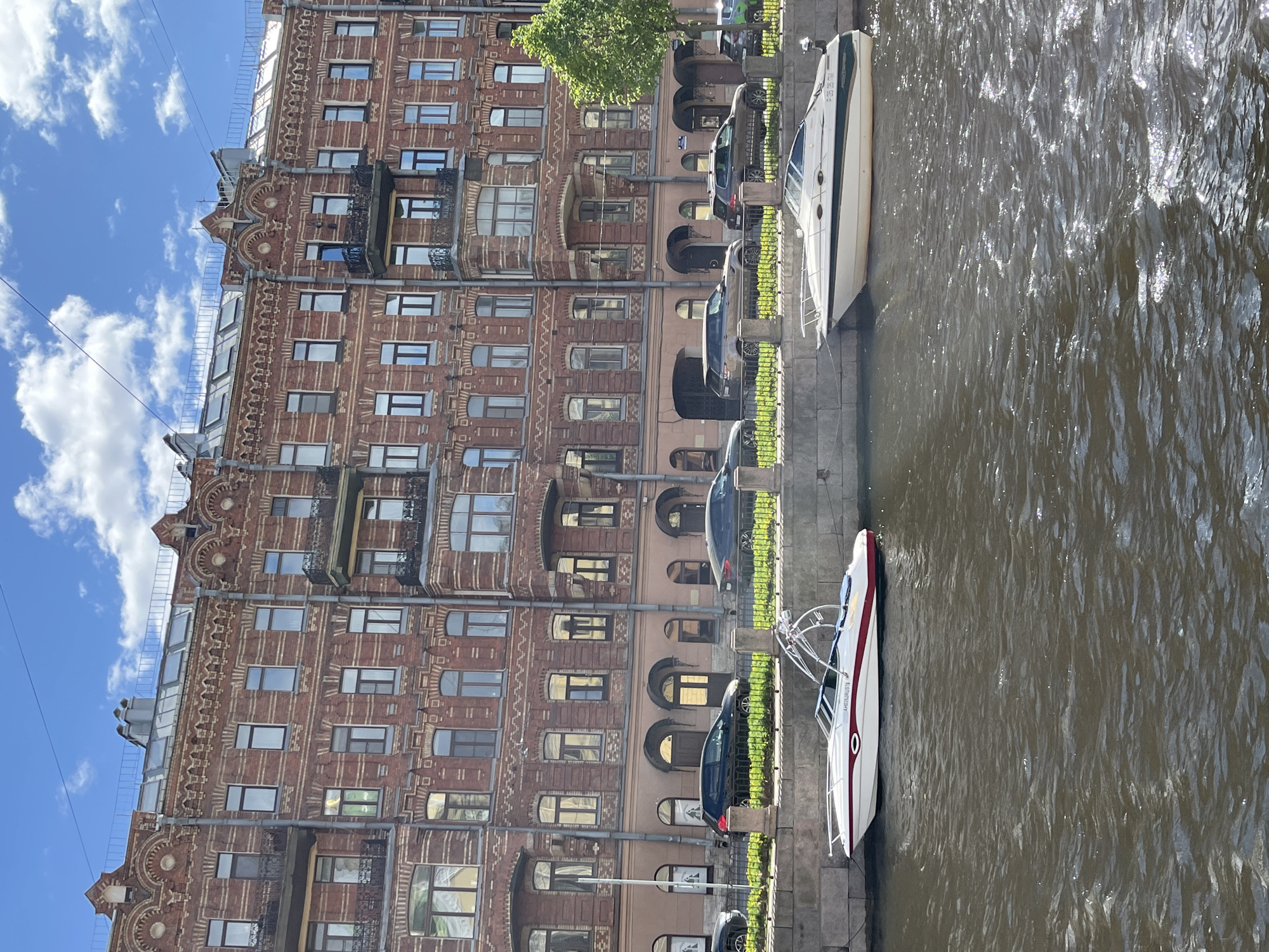 The image captures a canal scene in Saint Petersburg, Russia, with a detailed brick building on the left. The building has ornate architectural features, including decorative molding, balconies, and numerous windows reflecting sunlight. Several cars are parked along the canal, with some obscured by foliage. Two boats are docked on the canal's edge; one is white with red and blue trim, and the other is predominantly white. The canal water is rippled, reflecting light. The sky shows a few puffy white clouds against a blue backdrop.